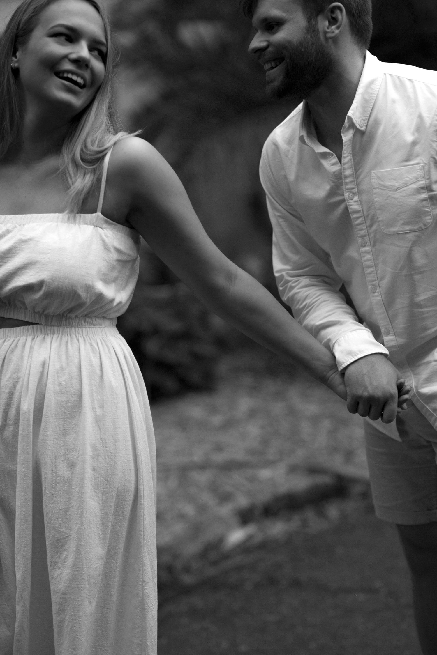 A couple shares a joyful moment outdoors in black and white, wearing casual white summer attire and holding hands.