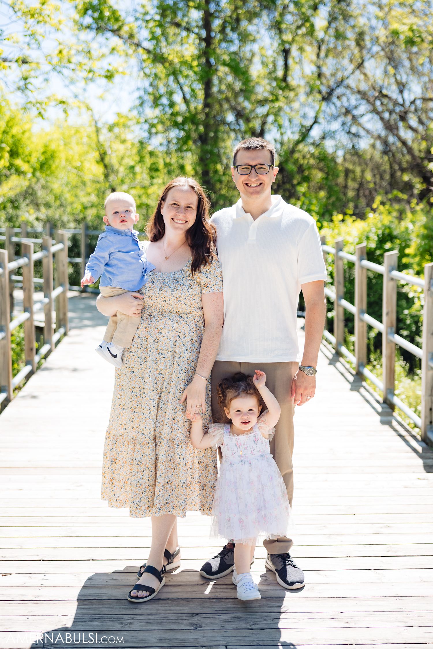 A family poses together on a white wooden boardwalk surrounded by green foliage on a sunny day.