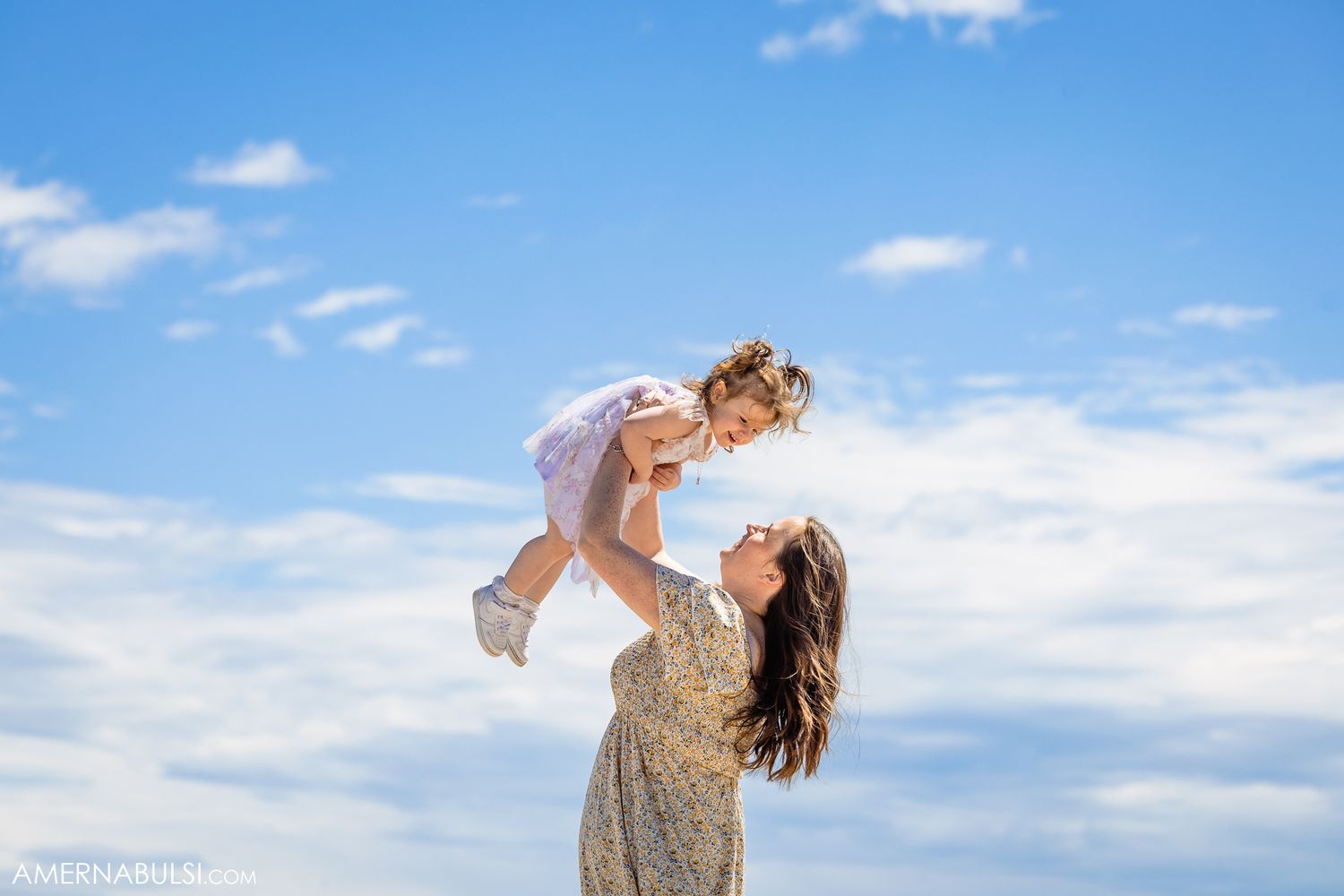A joyful outdoor moment captured against blue skies as a woman lifts a small child in the air.