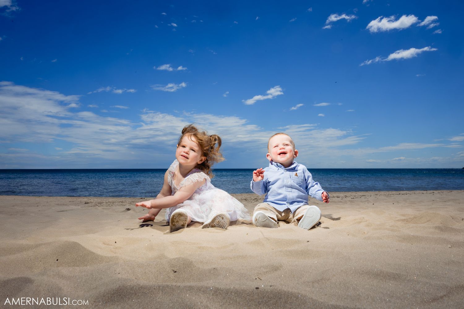 Two young children laughing while sitting together on a sandy beach with blue ocean and sky in the background.