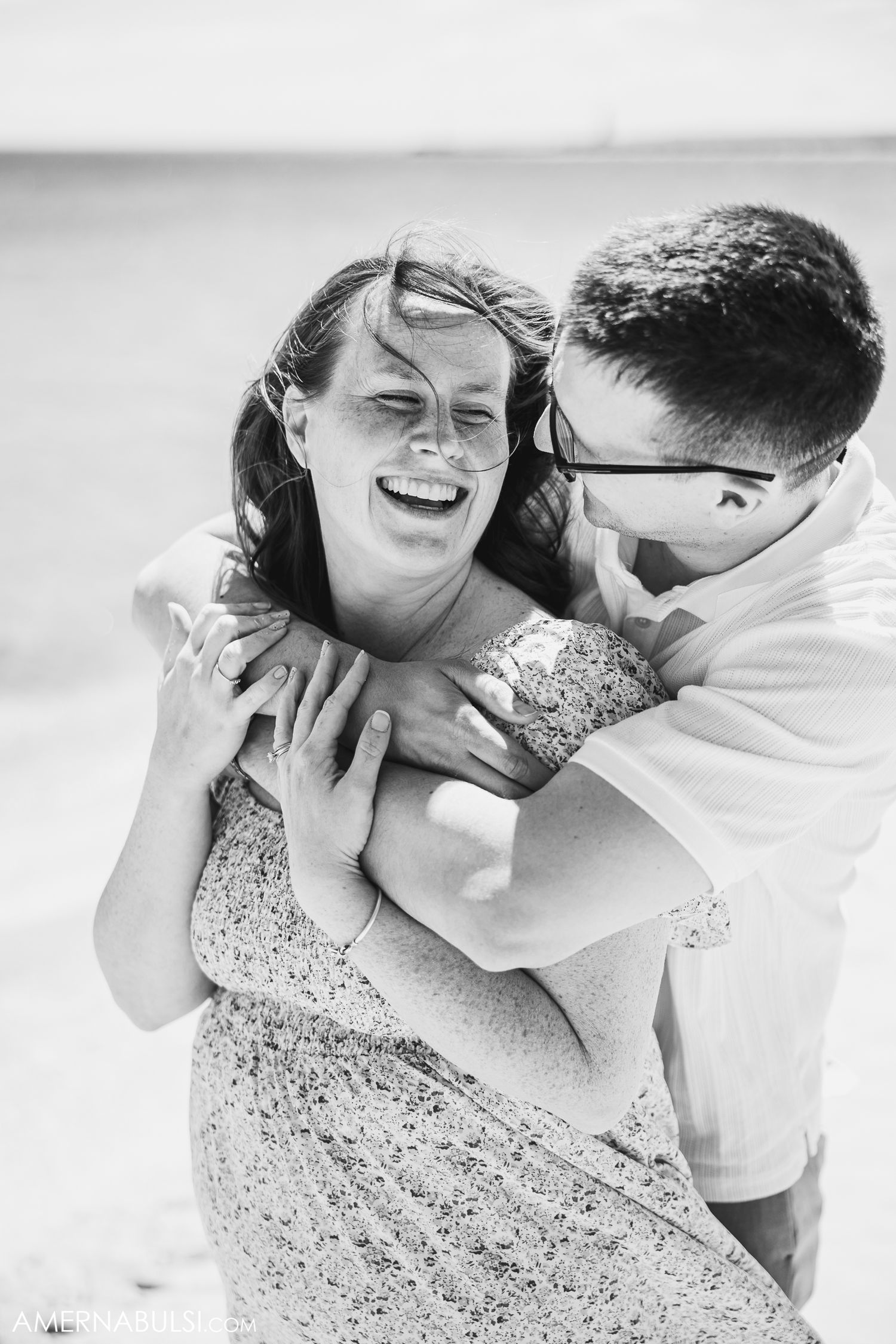 A candid black and white moment showing a couple sharing laughter and an intimate embrace against a bright background.