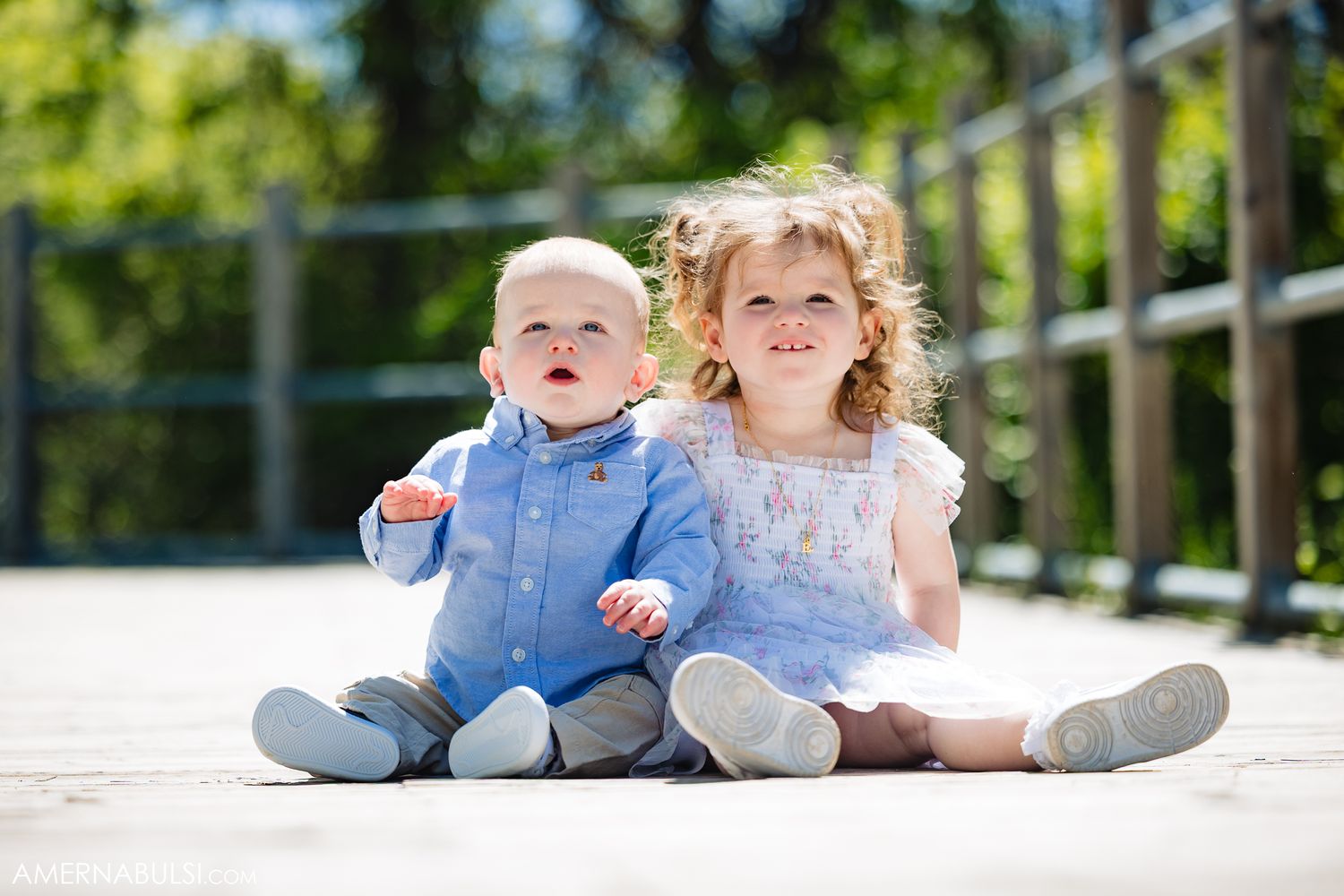 Two young children in formal attire sitting together and smiling outdoors on a bright sunny day.