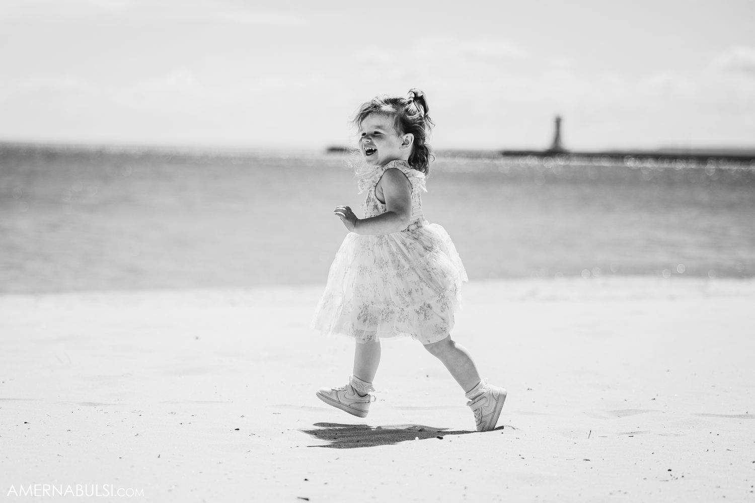 Black and white photo of a child running joyfully along the beach in a white dress.