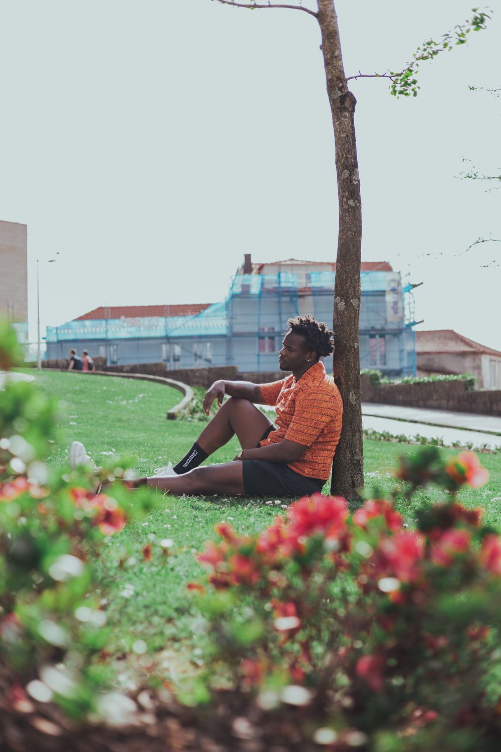 Person in orange shirt relaxing on grass near flowers with buildings in background.