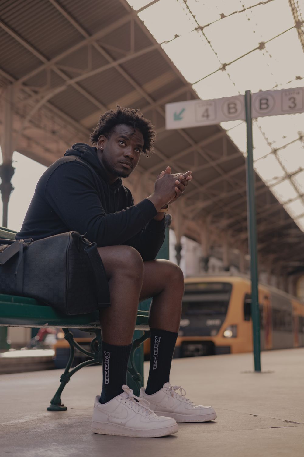 Person in dark hoodie sitting at train station platform with white sneakers.