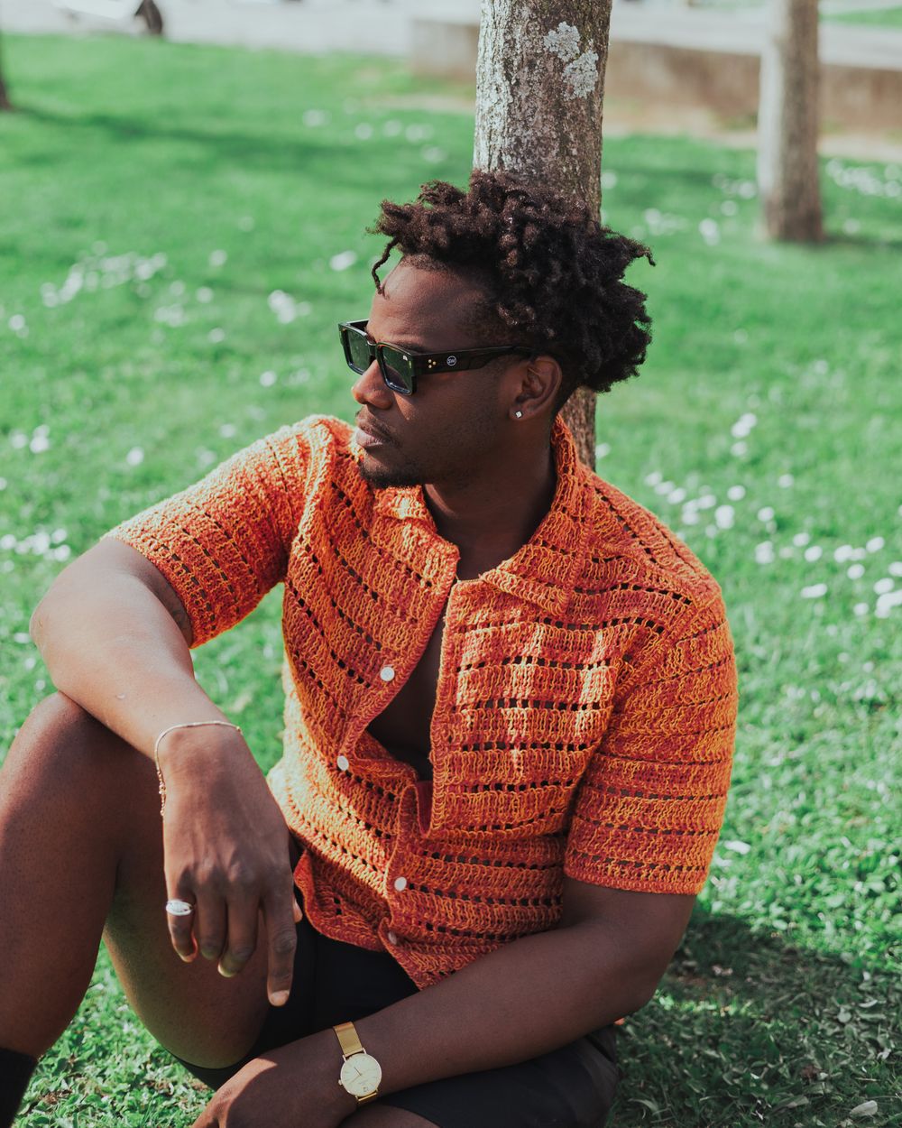 Person relaxing on grass wearing orange knit shirt, sunglasses and watch on sunny day.