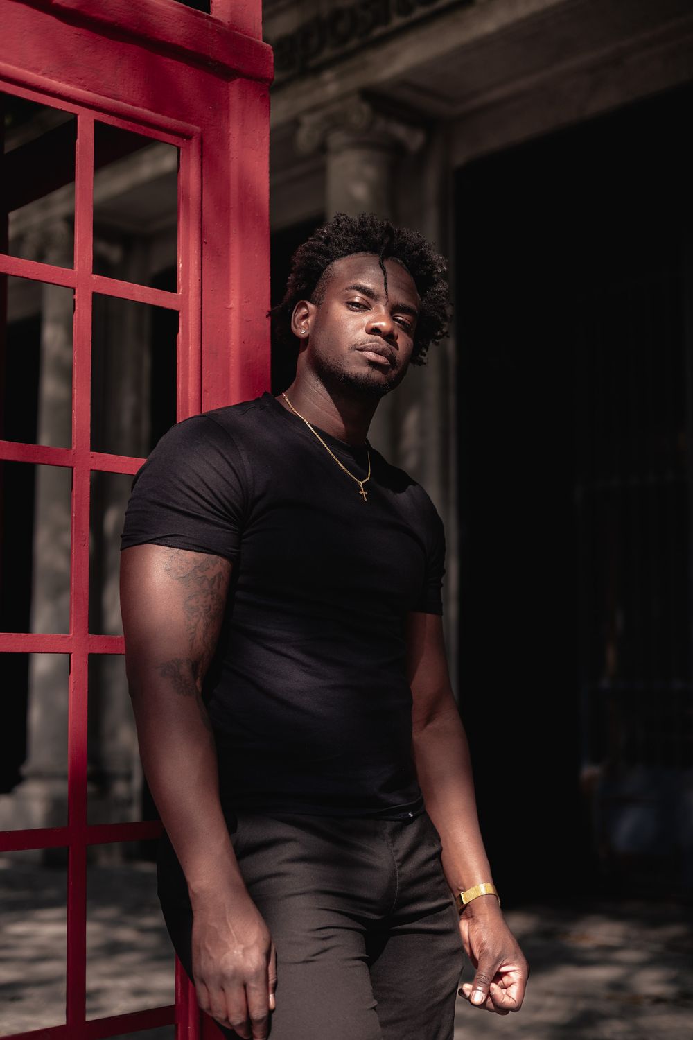 Dramatic portrait next to red phone booth in fitted black t-shirt and gold necklace.