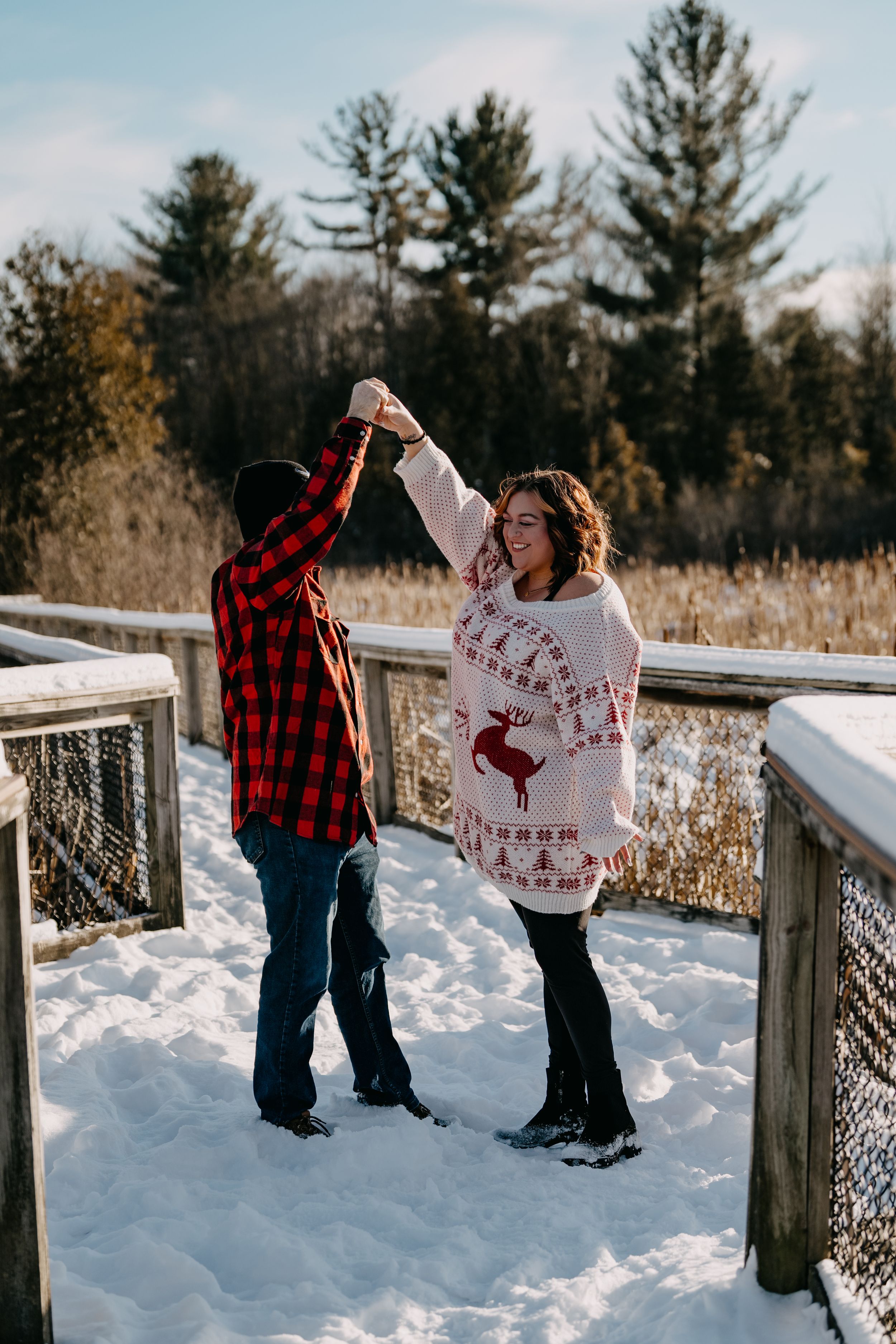 A couple dances playfully on a snowy wooden deck surrounded by evergreen trees on a bright winter day.