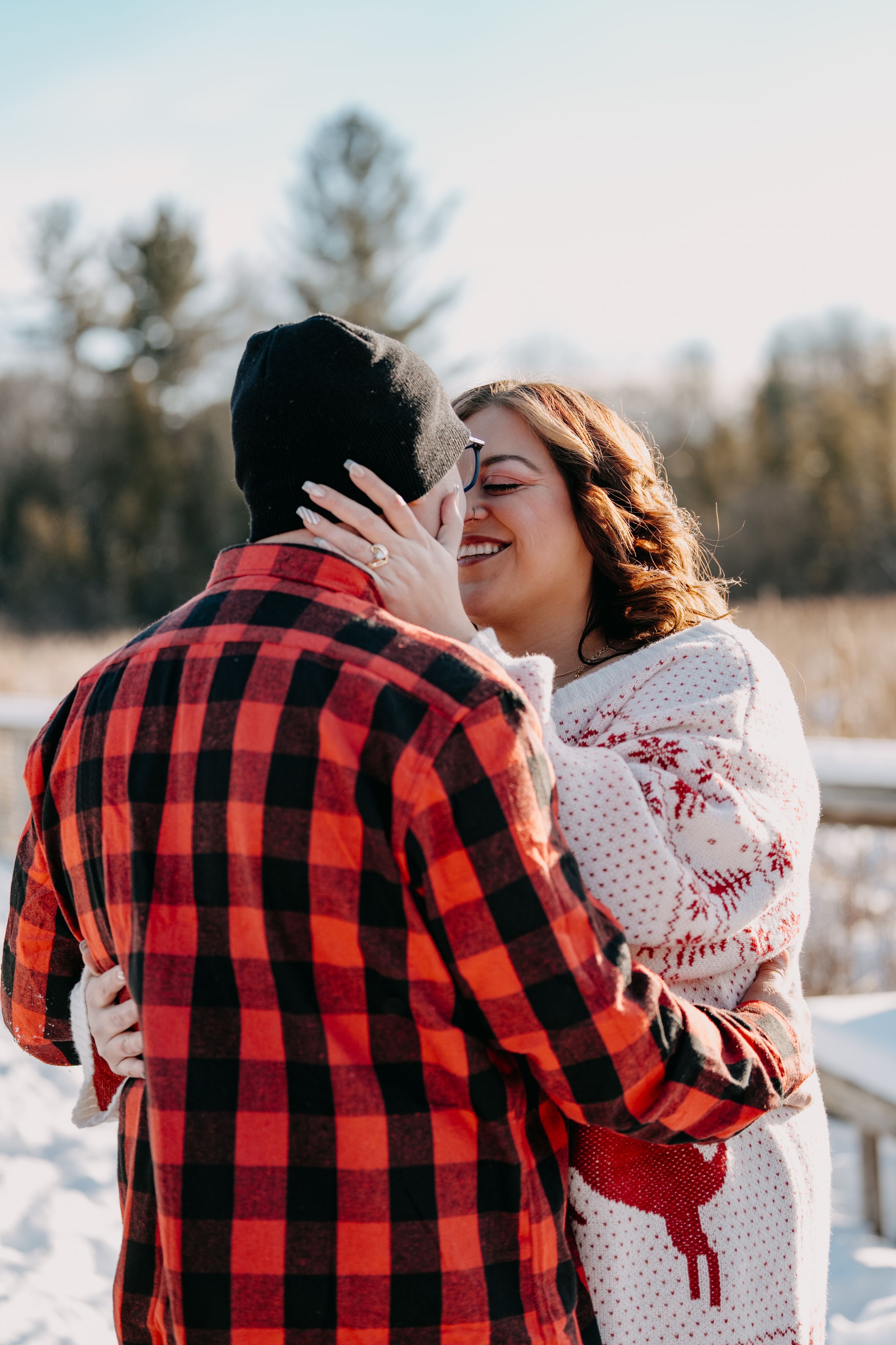 Couple embraces in winter scene with person wearing buffalo plaid flannel and other in festive reindeer sweater.