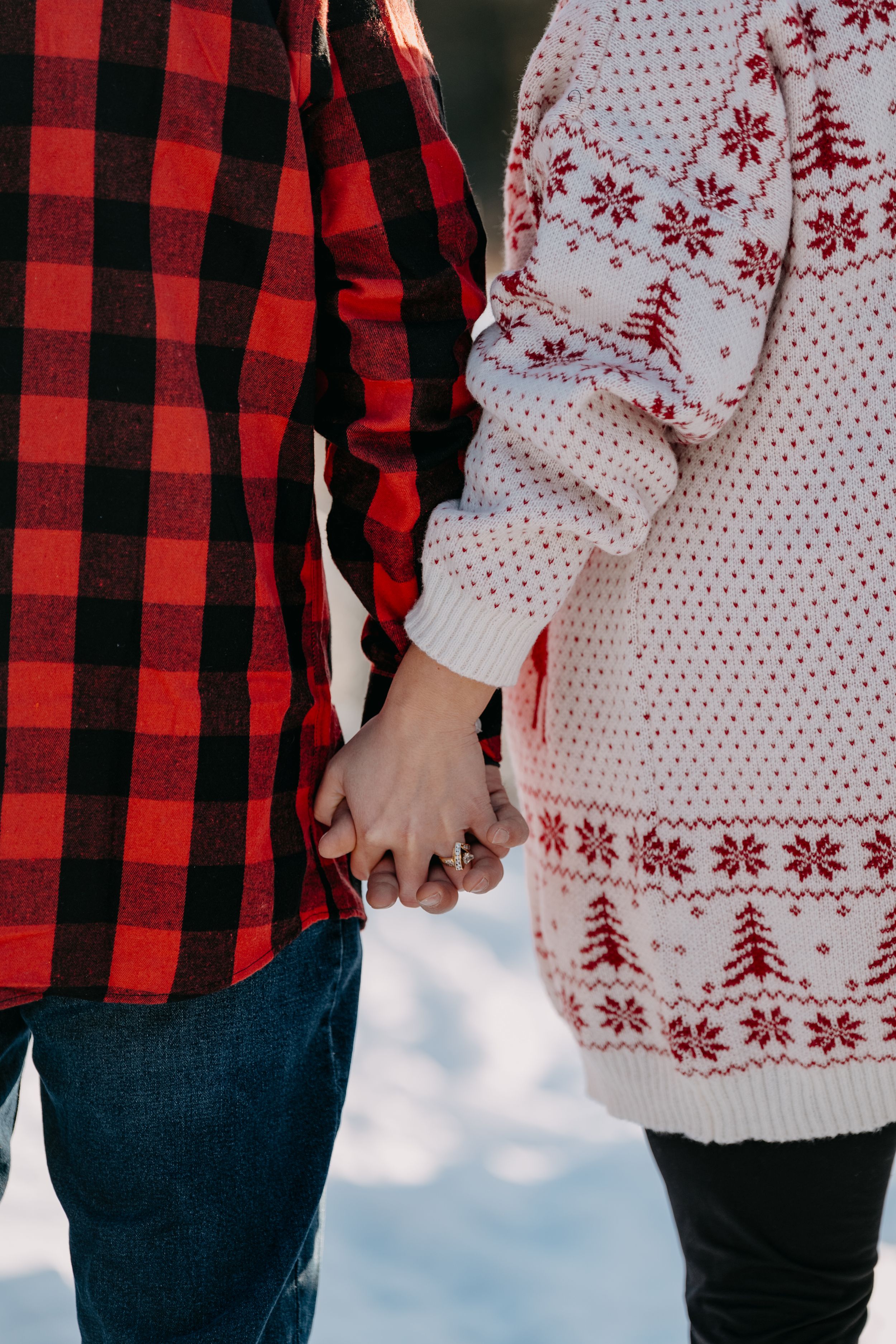 Couple holding hands wearing contrasting buffalo plaid shirt and white winter sweater with Nordic pattern against snowy backdrop.