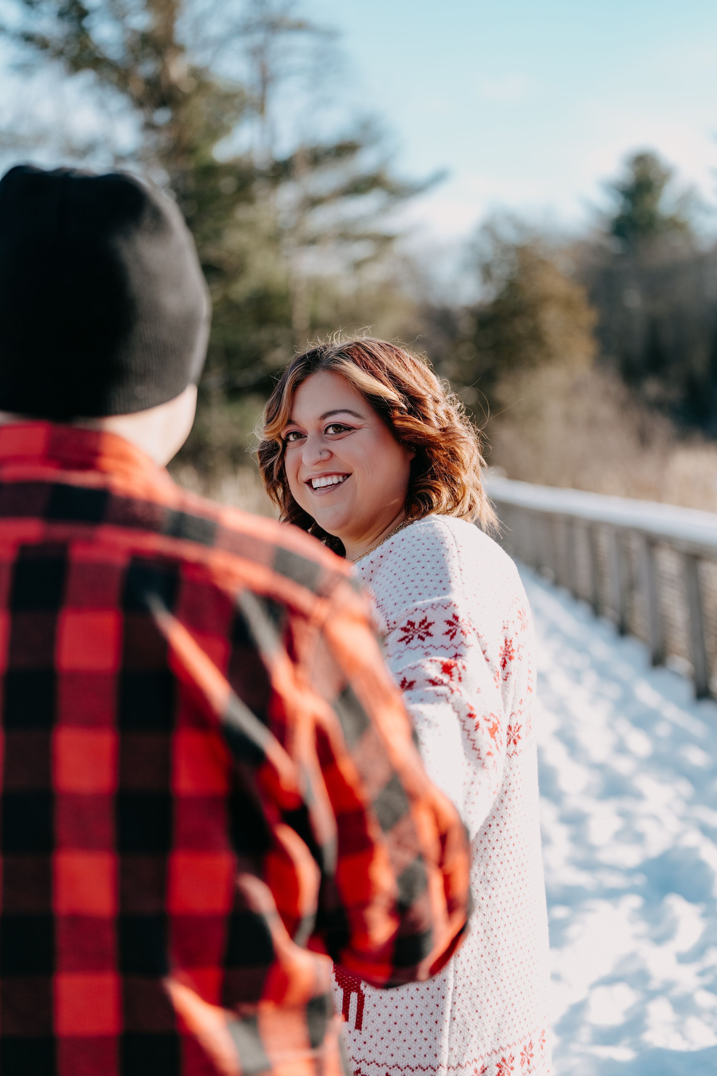 A person wearing a buffalo plaid flannel talks with someone in a white knit sweater on a snow-covered path.