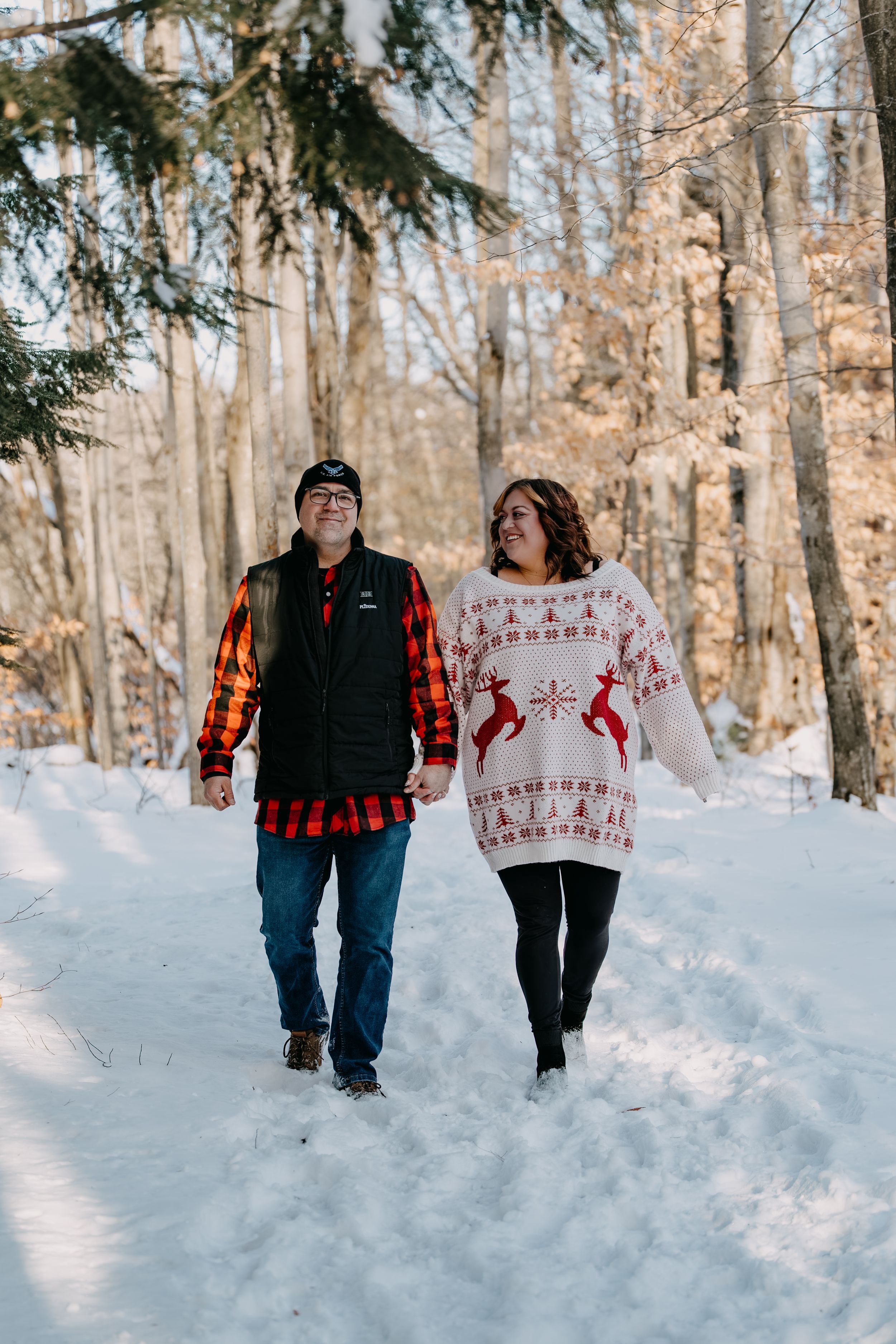 A couple walks together on a snowy winter path through the woods, wearing festive holiday sweaters and casual attire.