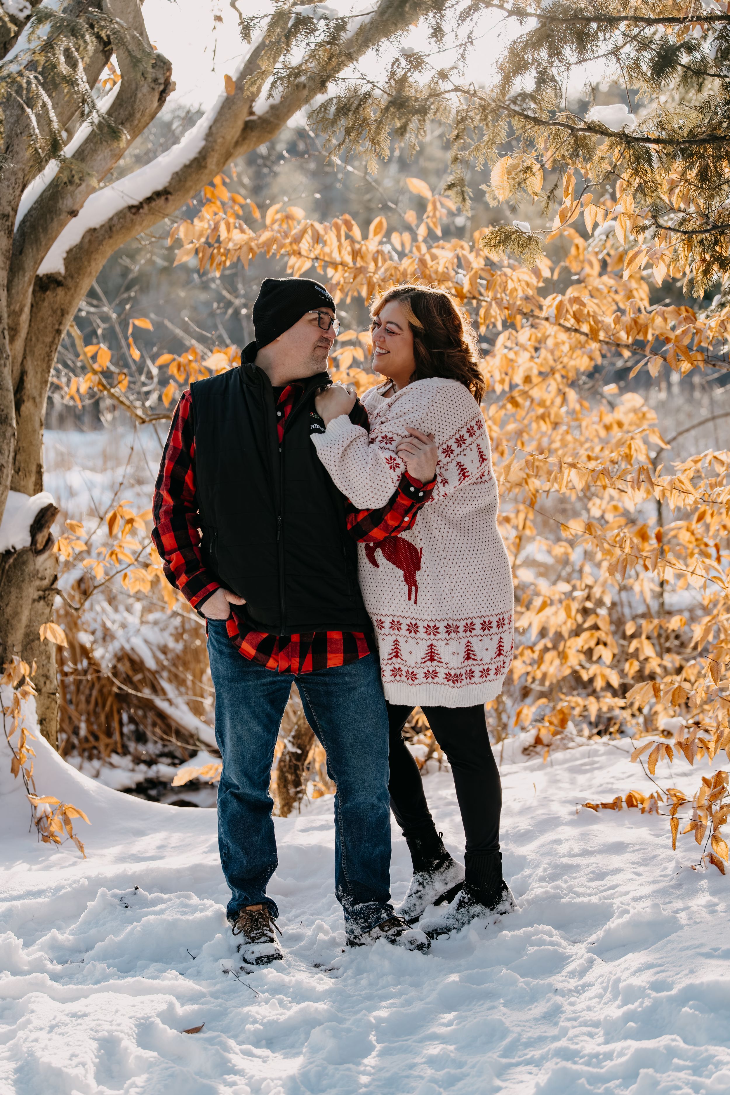A couple in winter clothing stands together in the snow with golden fall foliage glowing behind them.