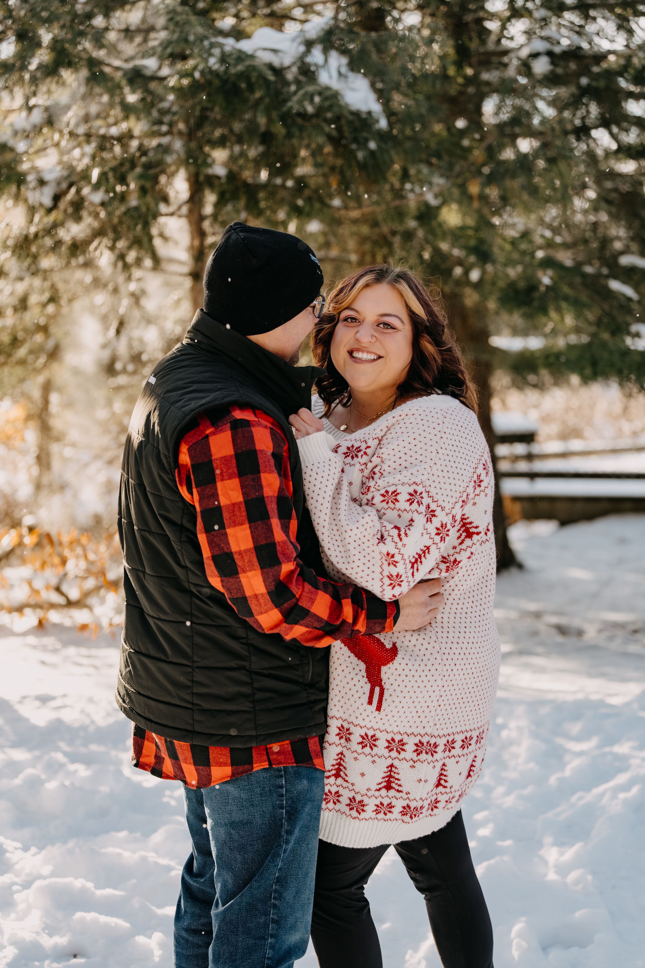 A couple embraces in a snowy winter scene with evergreen trees, wearing a buffalo plaid shirt and holiday sweater.