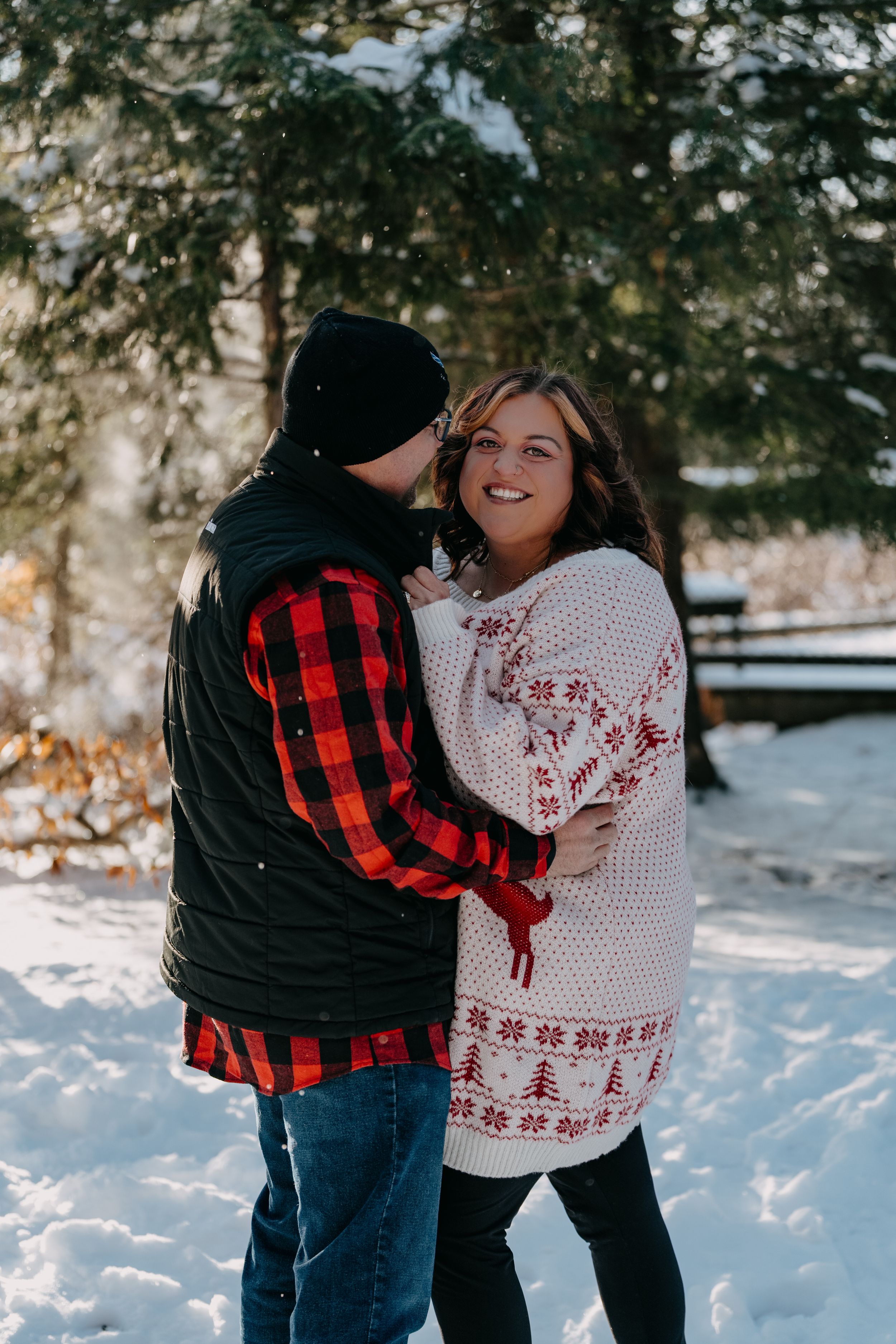 Couple embraces in snowy winter scene wearing red buffalo plaid and white knit sweater outdoors among evergreen trees.