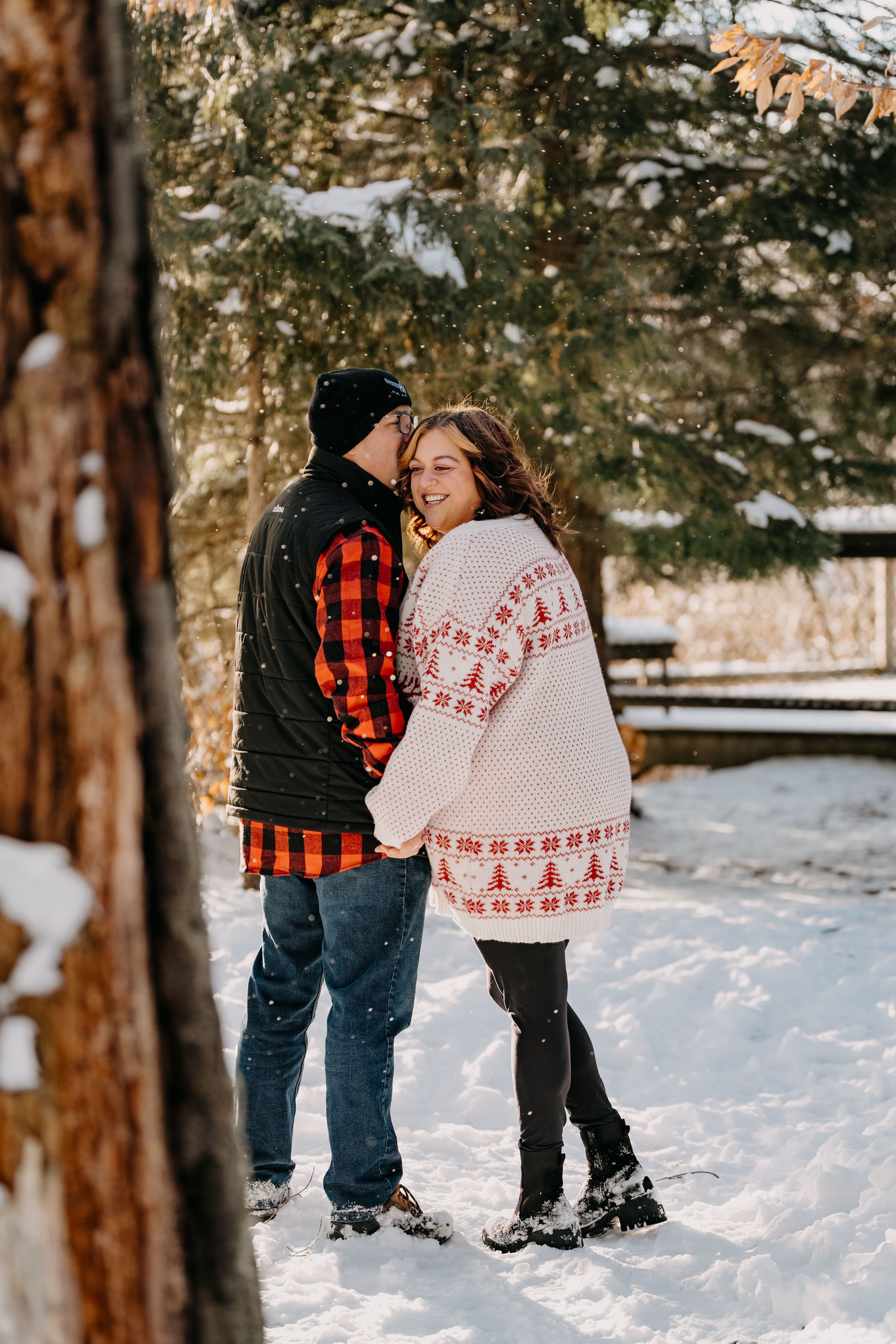 Couple in winter clothing sharing a romantic moment in snowy woods wearing plaid and festive knit sweaters.