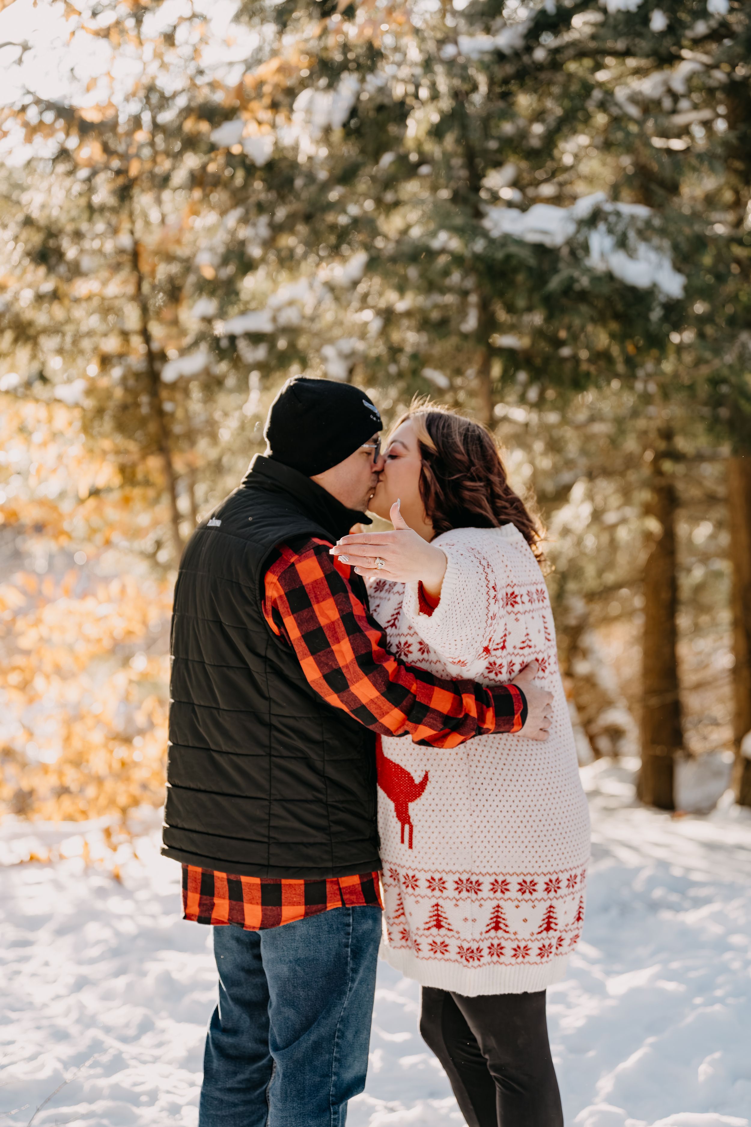 A couple embraces in a snowy forest wearing winter clothes, with one in a buffalo plaid shirt and one in a festive sweater.