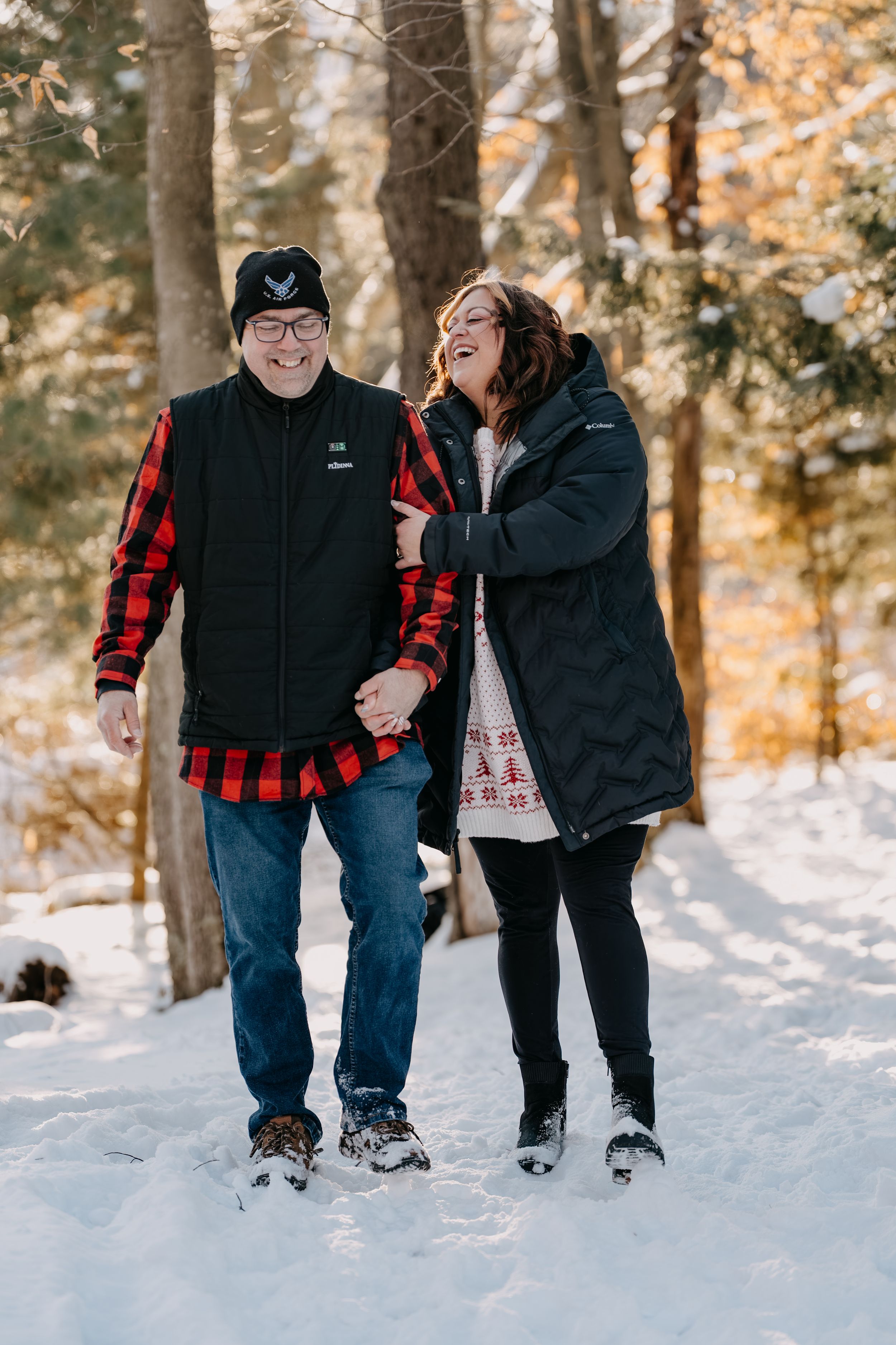 Happy couple laughing together while walking through snowy forest in matching buffalo plaid and winter gear.