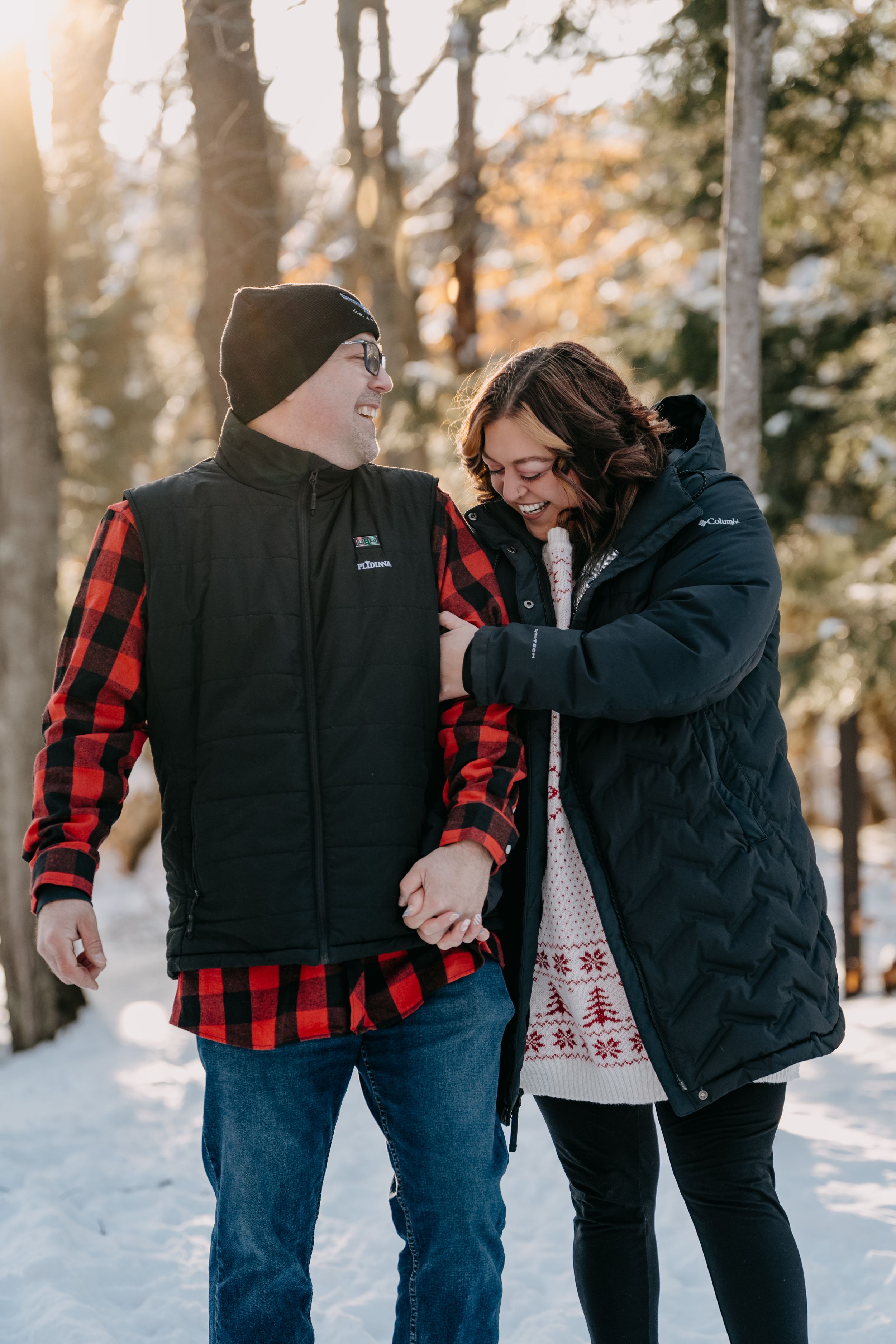 Two people in winter coats share a joyful moment together while walking in a snowy forest at sunset.