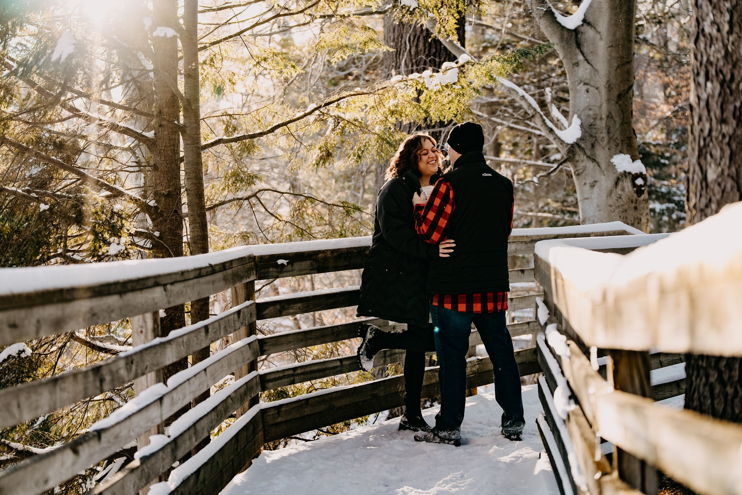 A couple in winter coats share an intimate moment on a snowy wooden bridge in a sunlit forest.