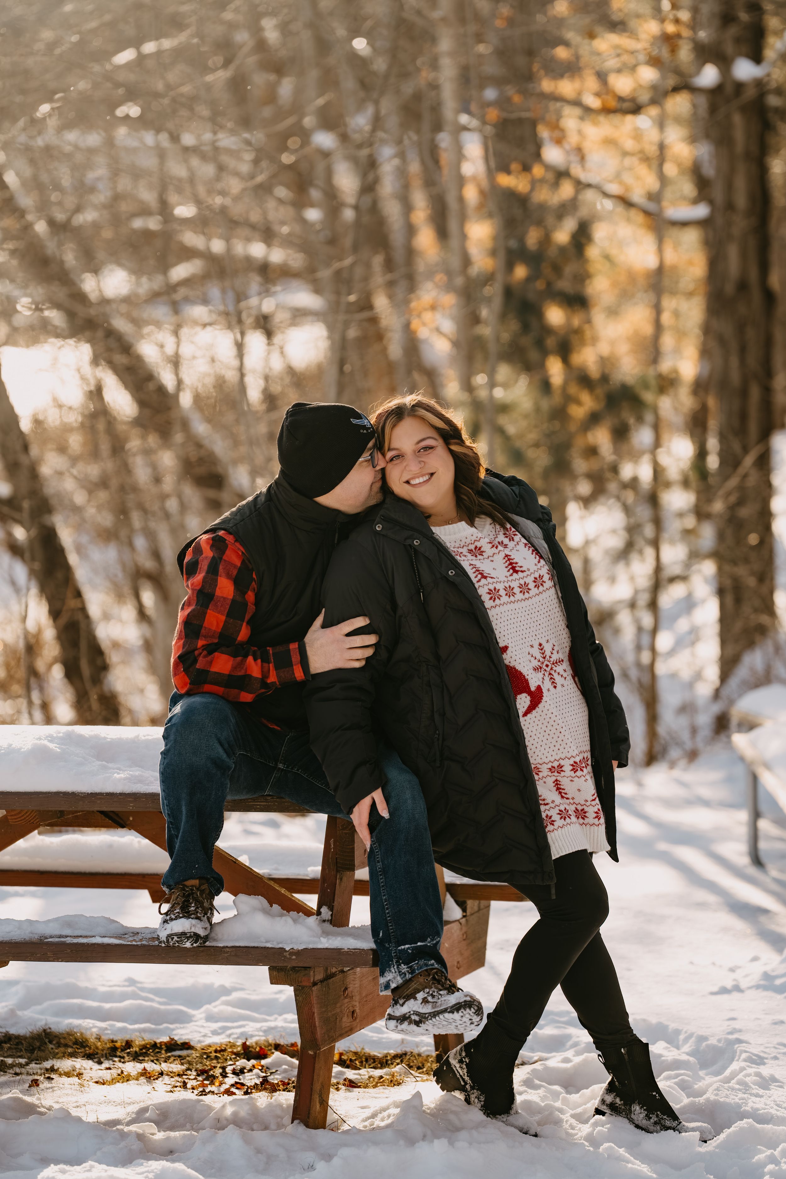 A couple shares an intimate moment on a wooden bench in a snowy winter forest scene with warm autumn colors in background.