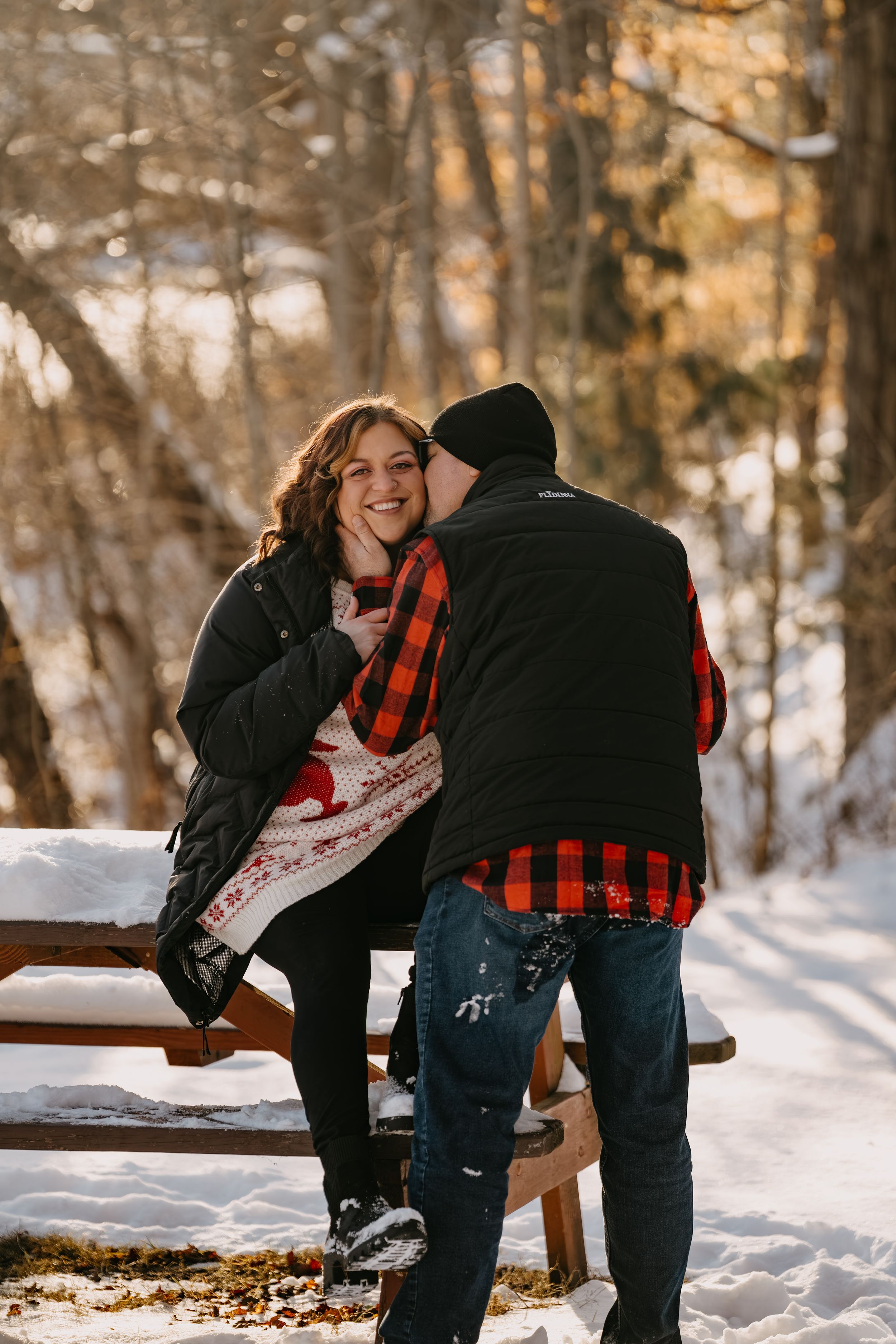 A couple shares a tender moment on a snowy wooden bench in a winter forest setting, wearing buffalo plaid and black jackets.