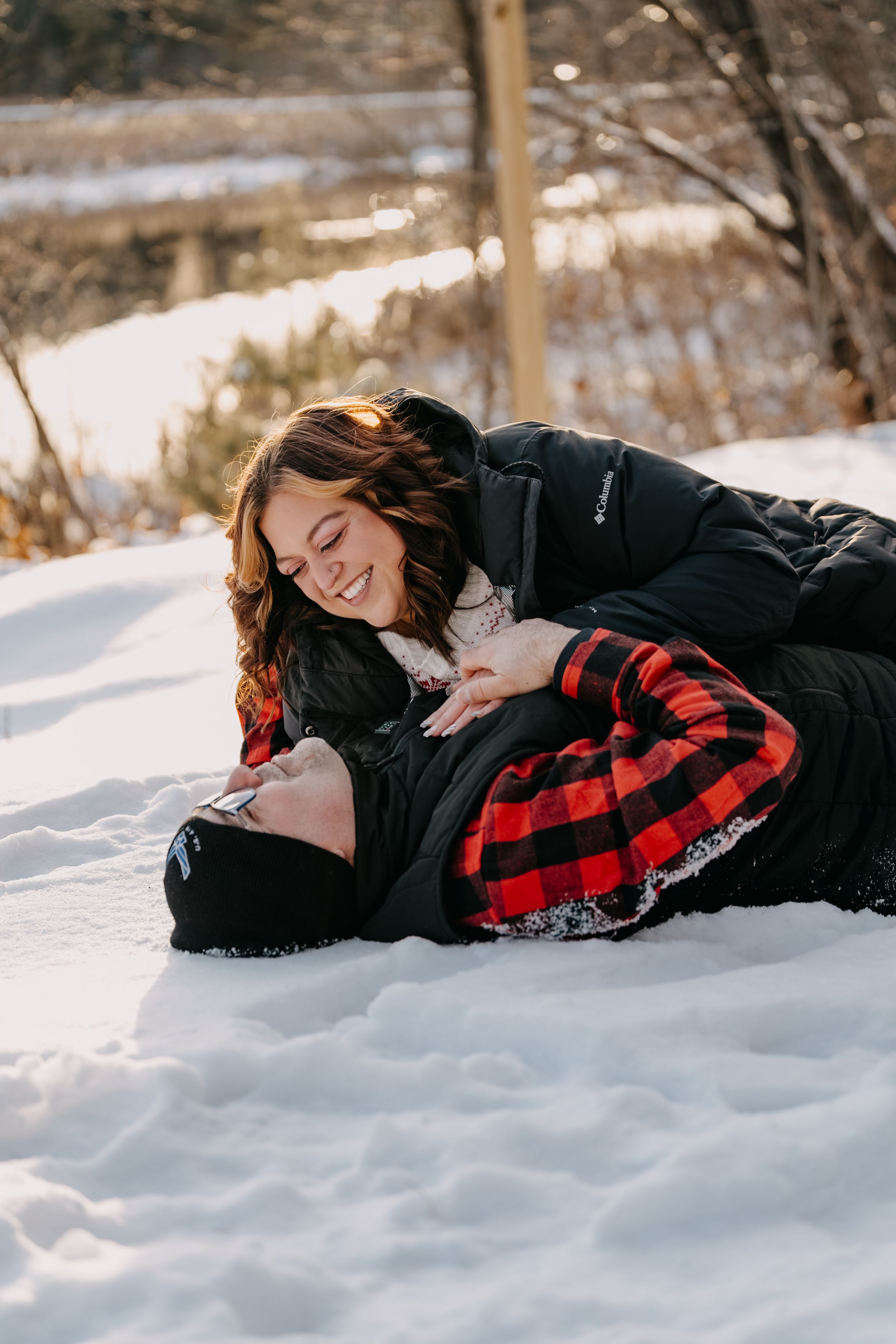 Woman in black coat and red plaid scarf lies peacefully in fresh white snow during winter photo session.