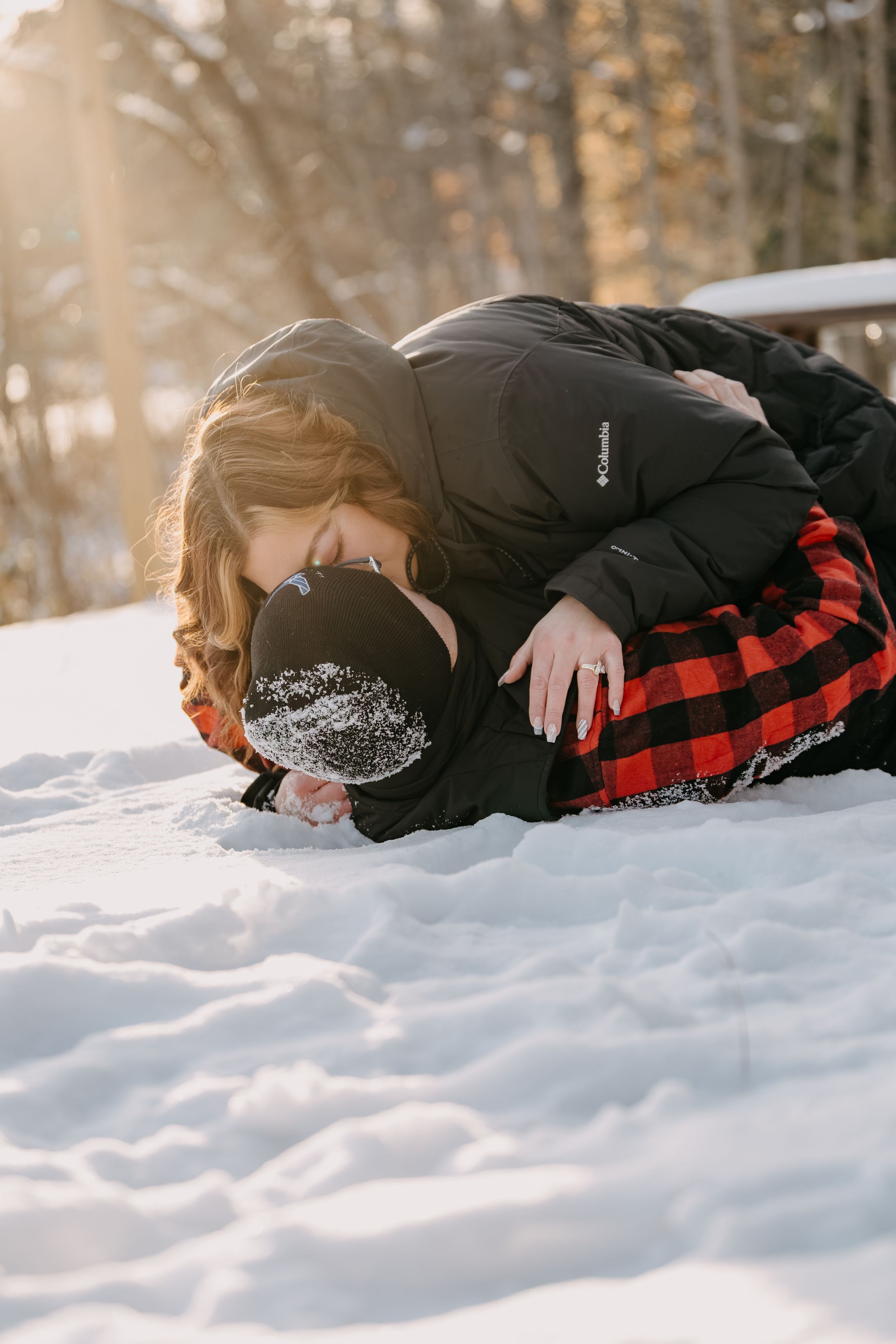 Someone in a plaid shirt and black coat lies face down in pristine white snow during a winter photo shoot.