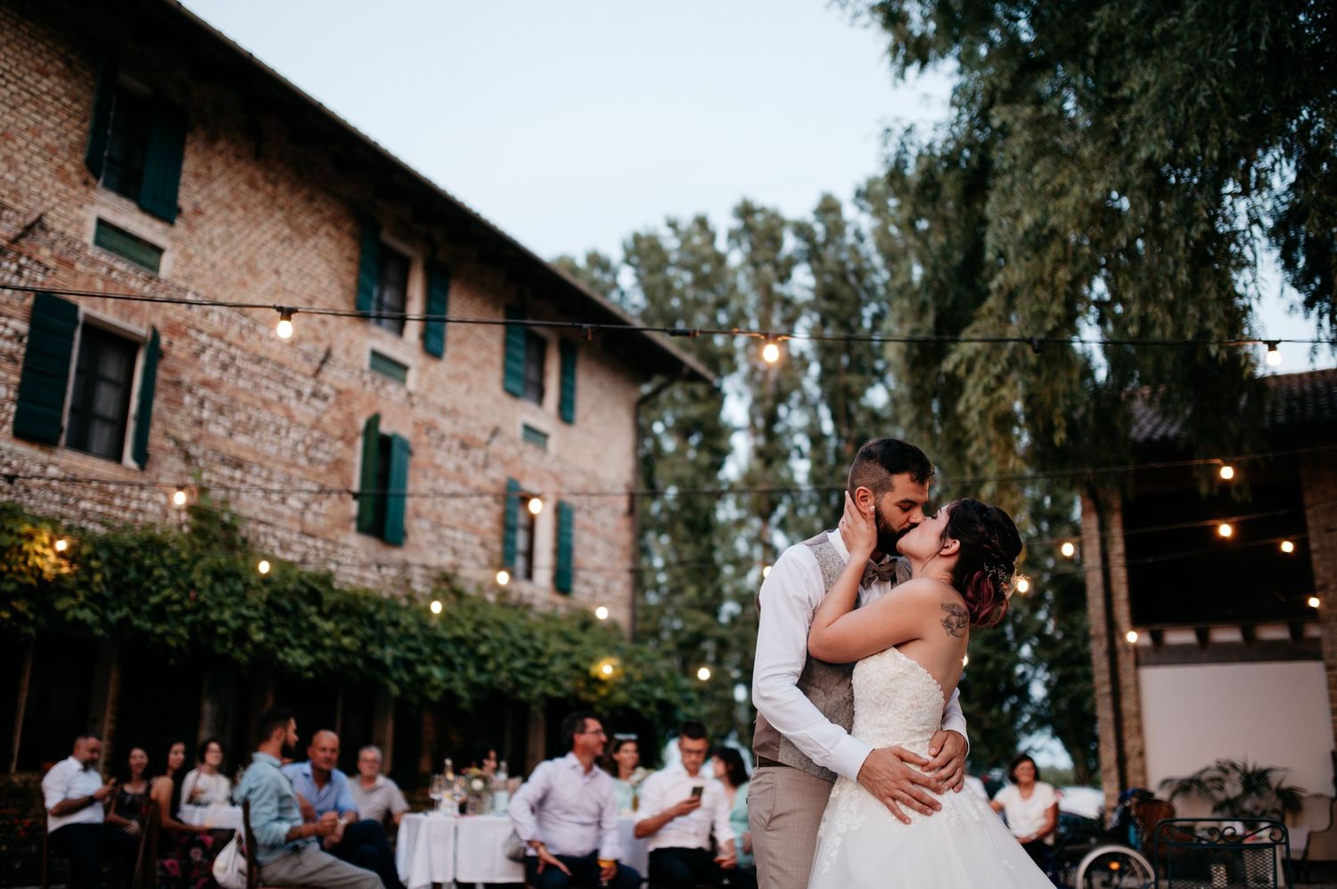 A romantic couple dances together during a sunset wedding reception at an outdoor venue with string lights overhead.