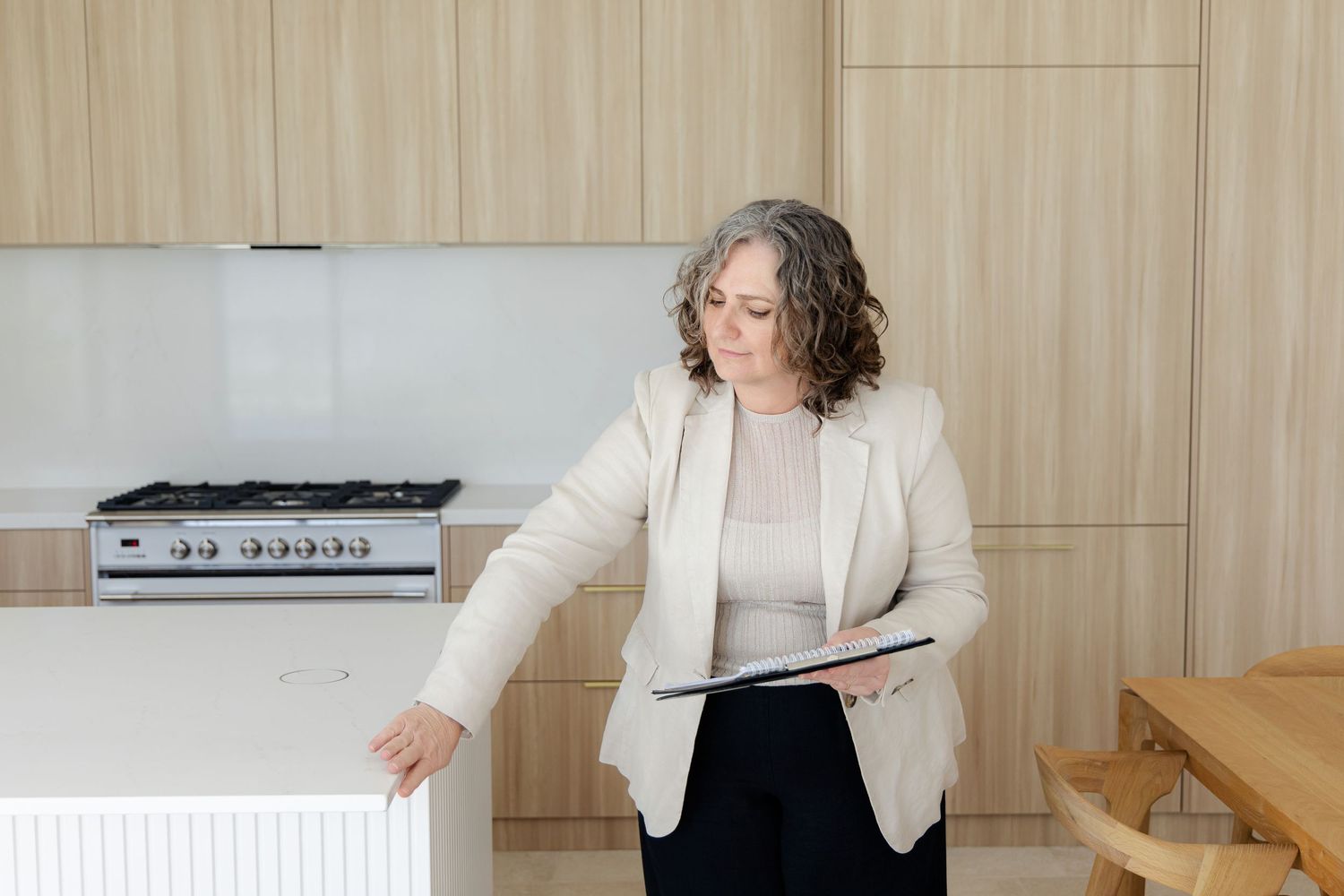 Professional realtor in cream blazer gestures toward radiator while holding digital tablet in modern minimalist kitchen.