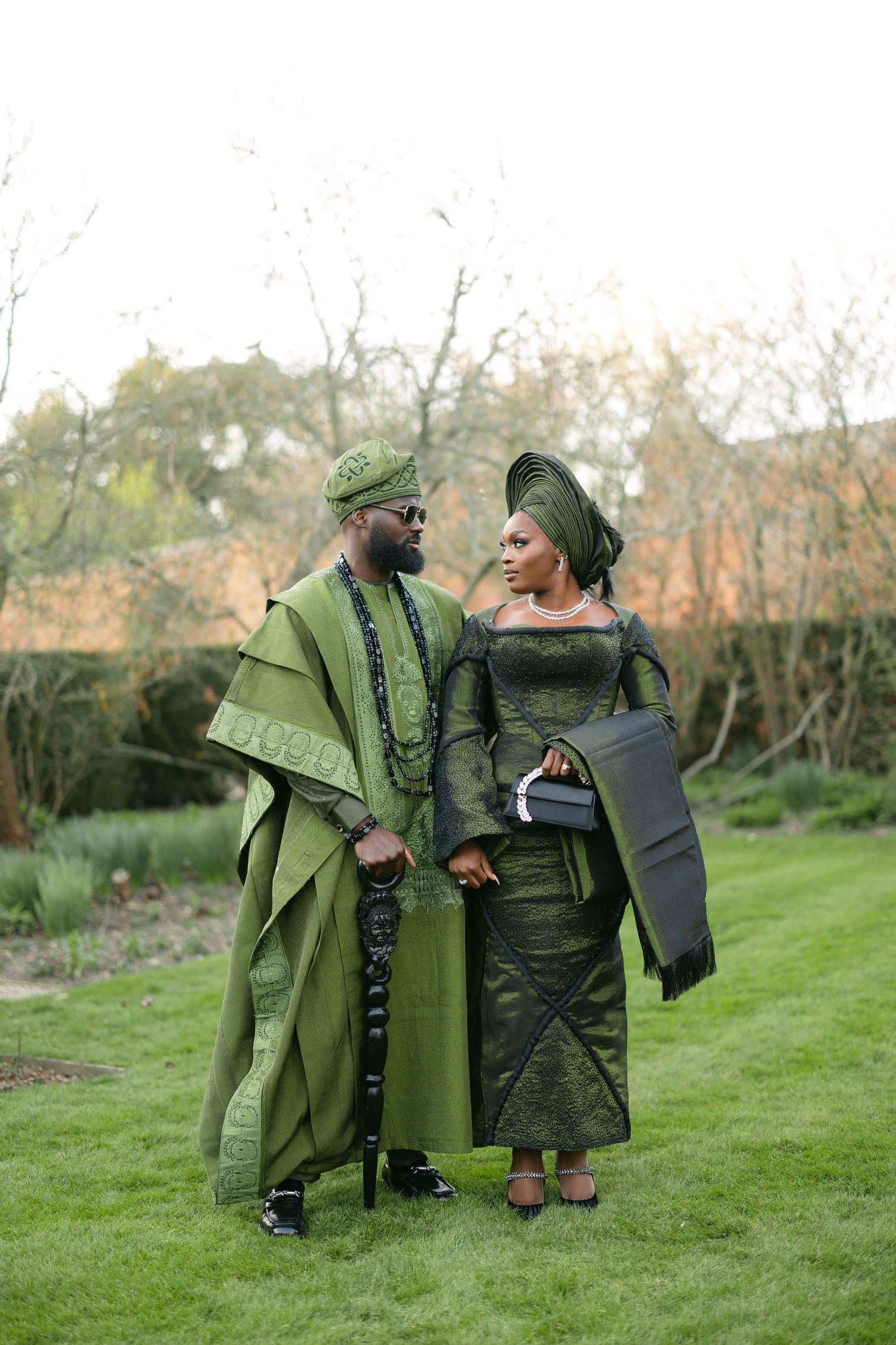 Couple in elegant green and grey traditional African attire pose together on grassy lawn during outdoor photoshoot.