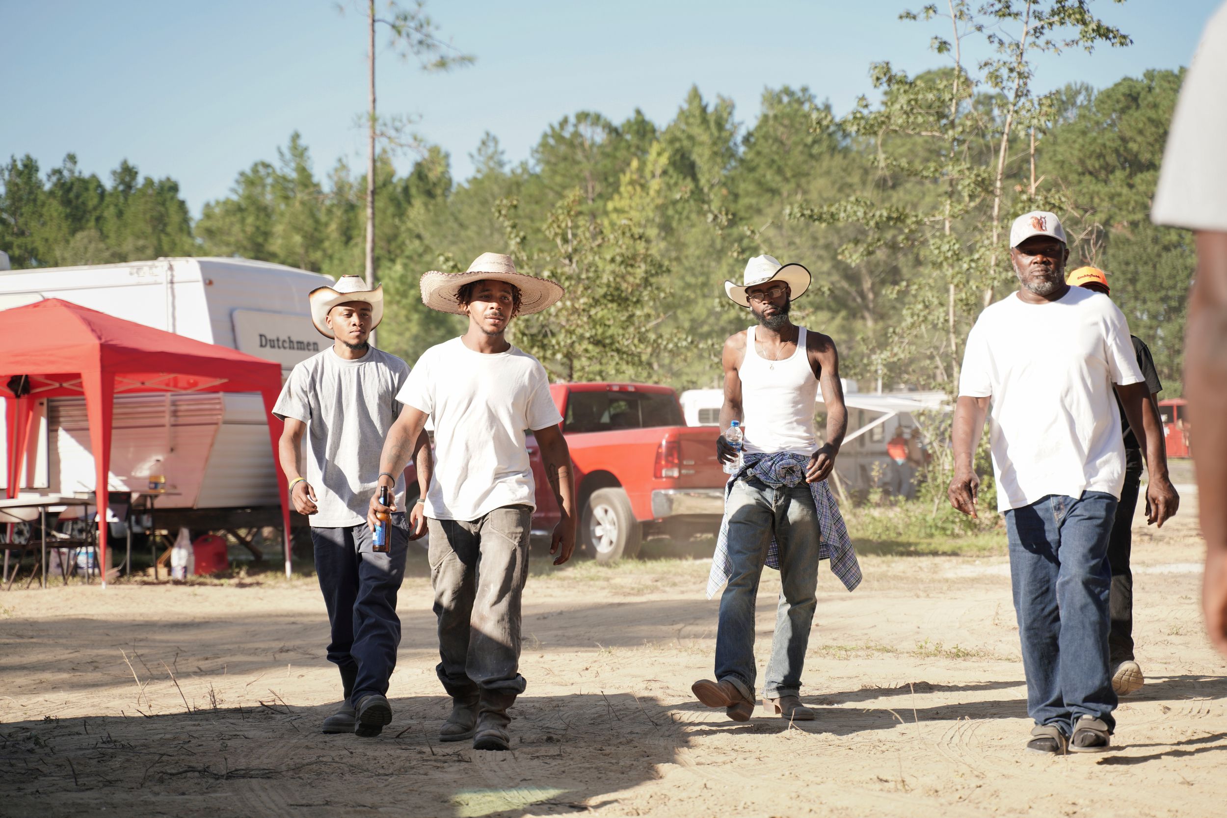 A panoramic view of workers gathered outdoors in a rural area near trees and red buildings on a sunny day.