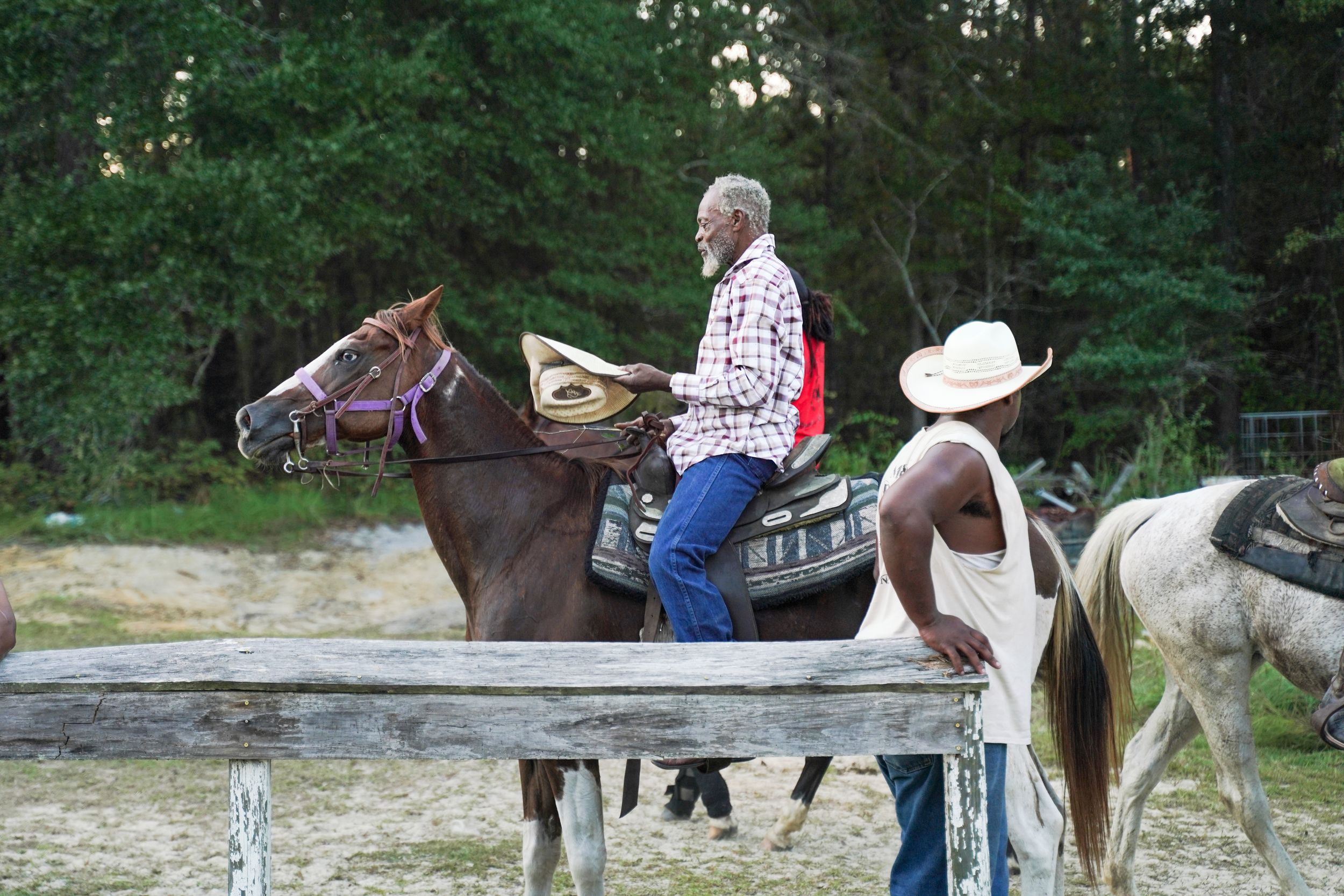 People on horseback gather at a wooden fence rail near a wooded area during an equestrian activity.