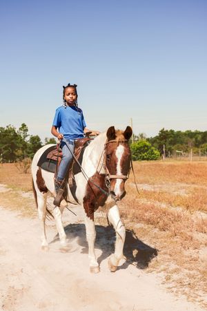A rider in blue on a brown and white paint horse trots across dusty ranch terrain on a sunny day.