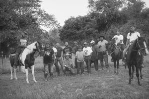 A vintage black and white photograph shows a group of people gathered with horses in a wooded outdoor setting.