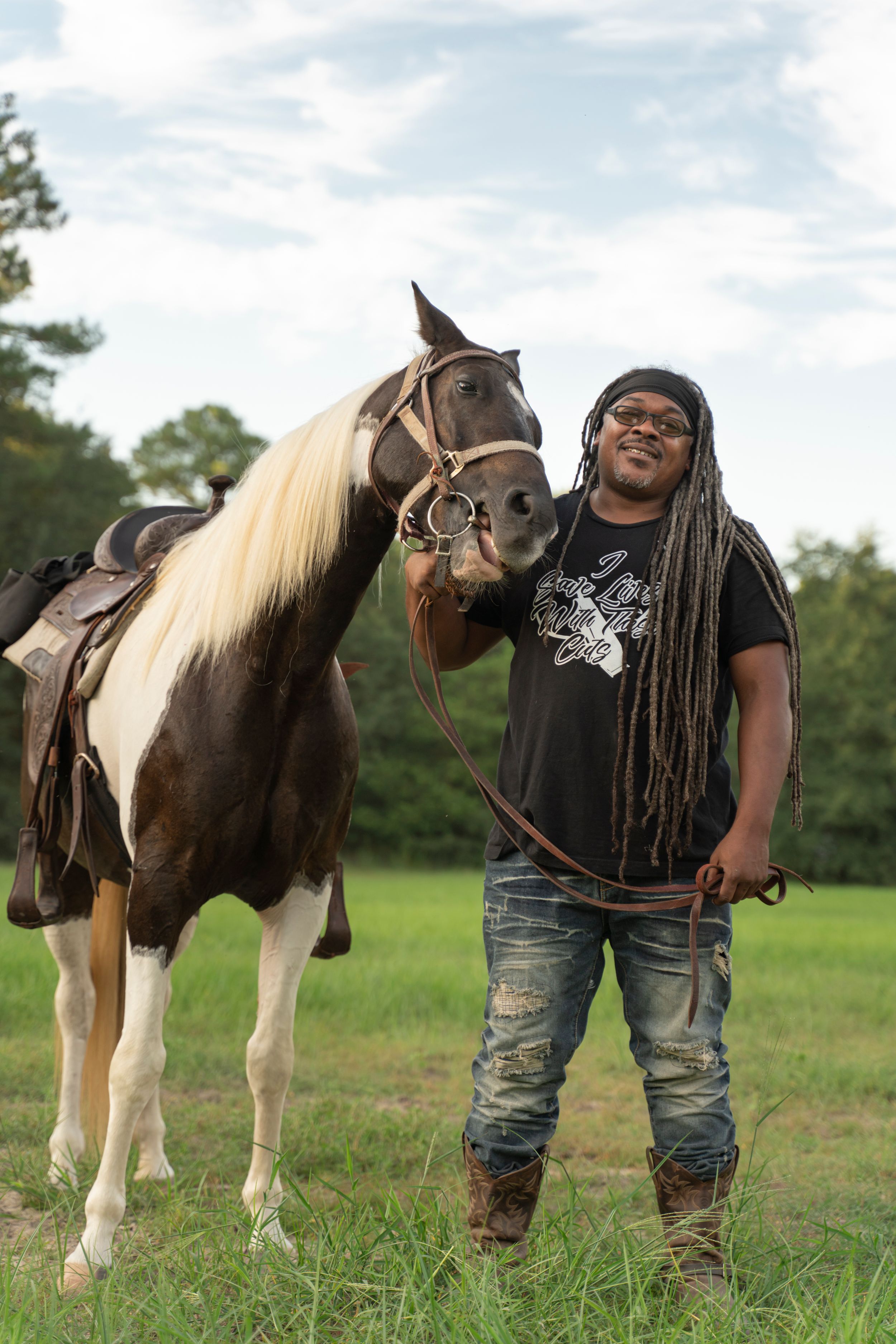 A paint horse with a brown and white coat stands in a grassy field next to a person wearing a black t-shirt and cowboy boots.