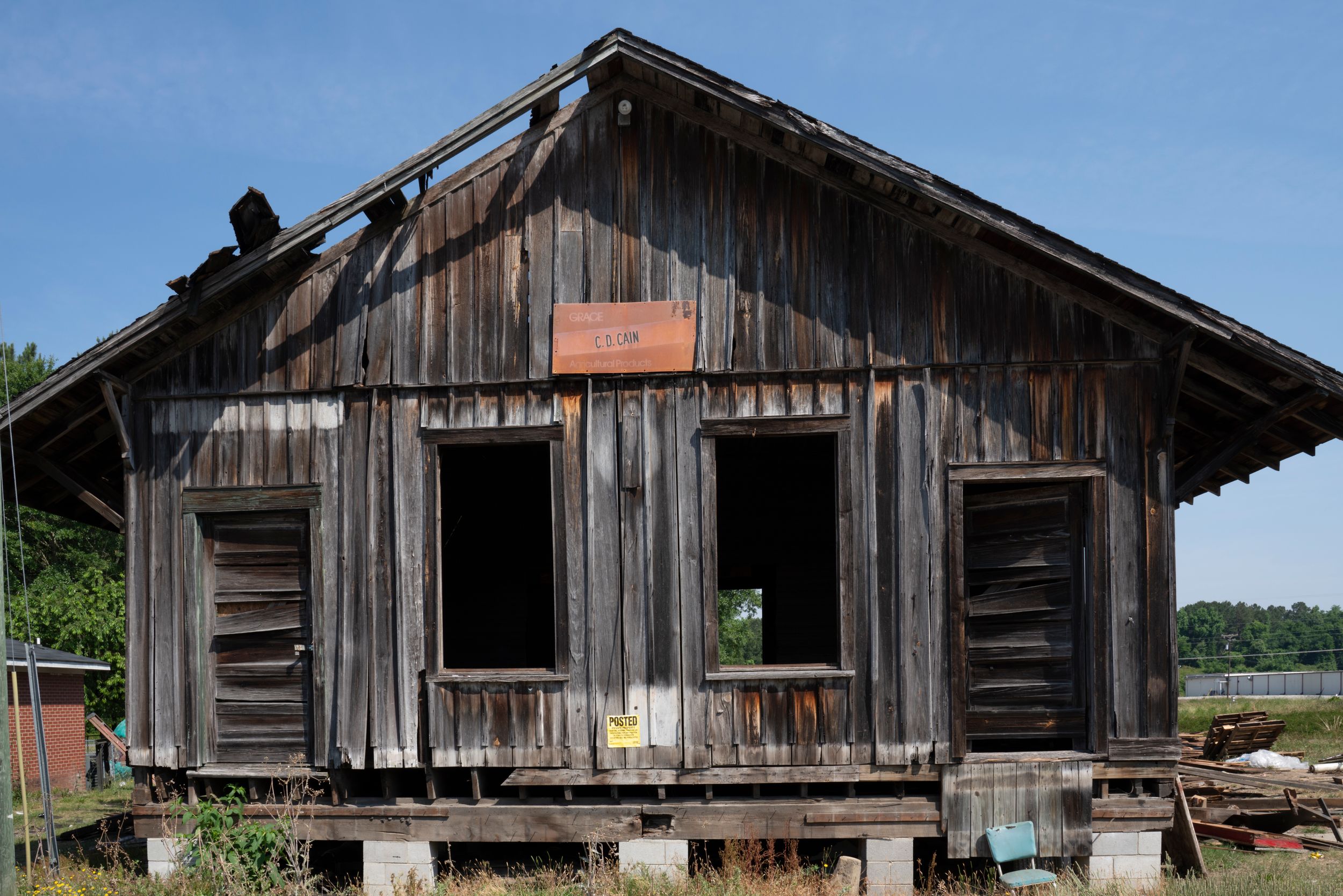 A weathered wooden shack building with broken windows and peeling boards sits abandoned in a rural setting.