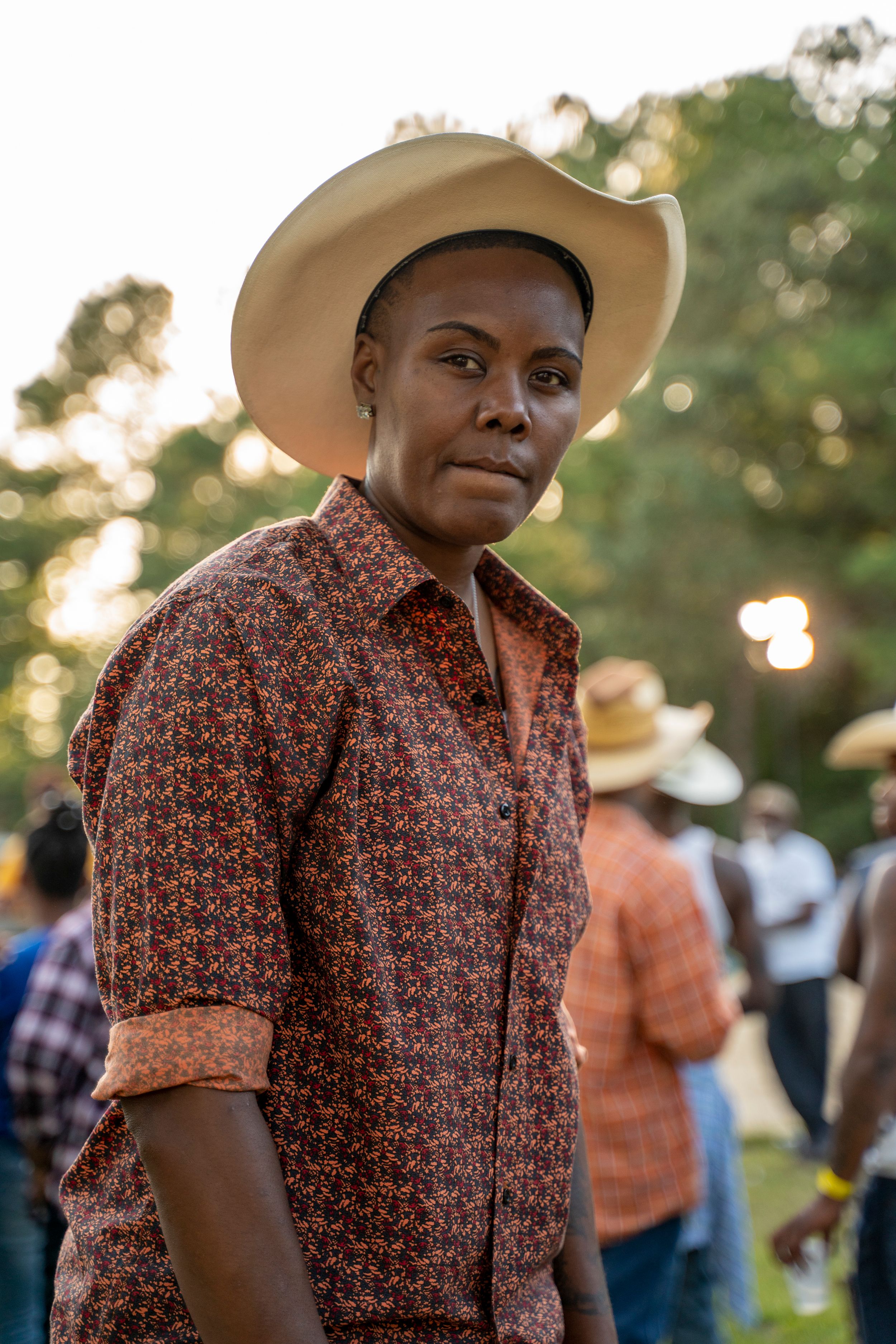 Black Cowboy in a cowboy hat and patterned button-up shirt stands outdoors during golden hour with a crowd in the background.
