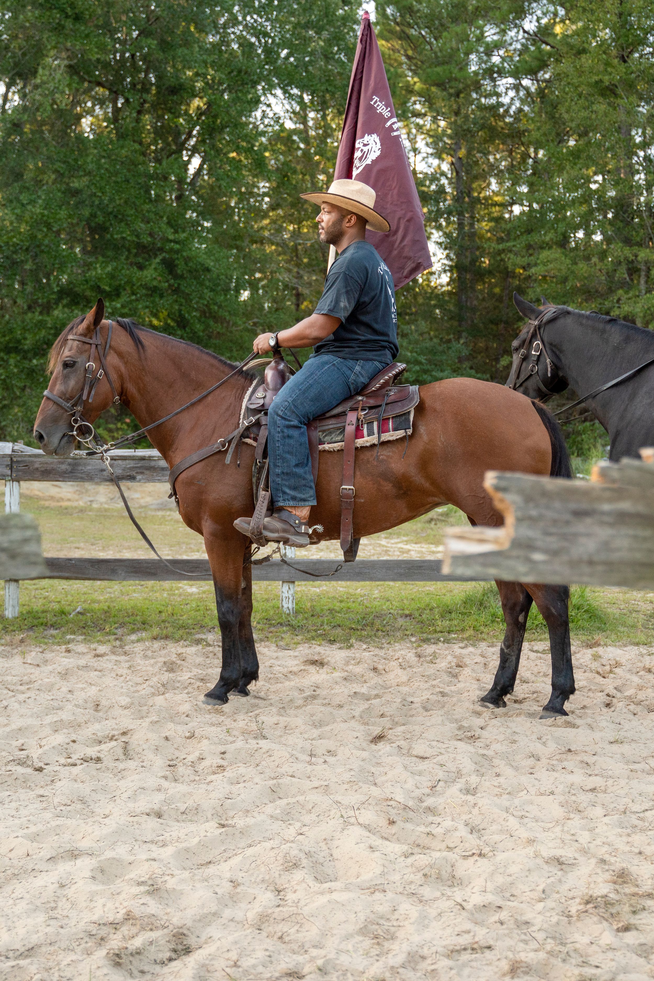 A rider mounted on a brown horse carrying a flag in a sandy arena with trees in the background.
