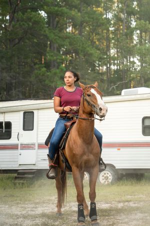 A person rides a chestnut brown horse near white trailers in a wooded area.