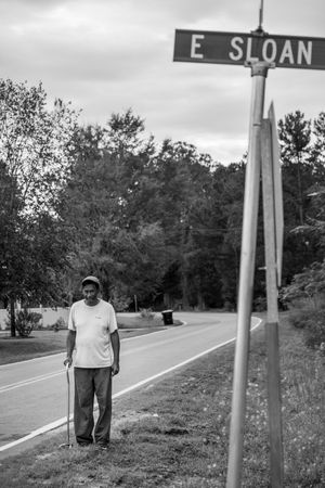 Black and white photograph of a street scene with a figure standing near an E Sloan street sign on a rural road.