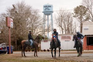 Three people on horseback gathered near a water tower in a small rural town setting during winter.