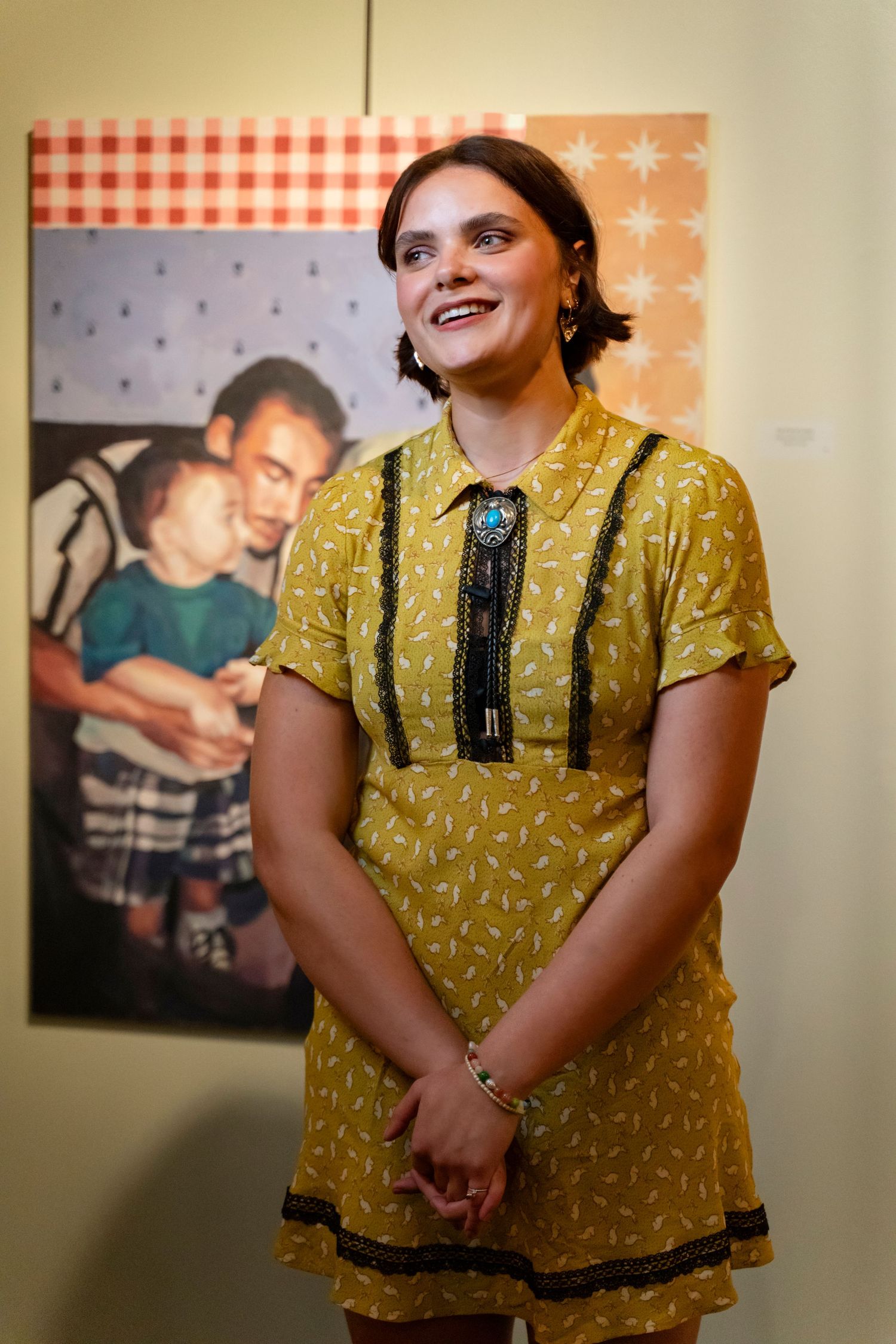 A person wearing a yellow patterned dress with black trim stands smiling in front of a framed photograph on a wall.