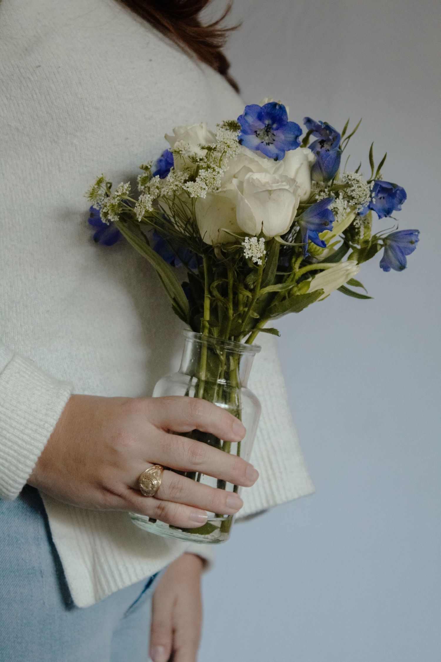 Hand wearing a white sweater holds a delicate bouquet of white roses and blue delphiniums in a glass vase.