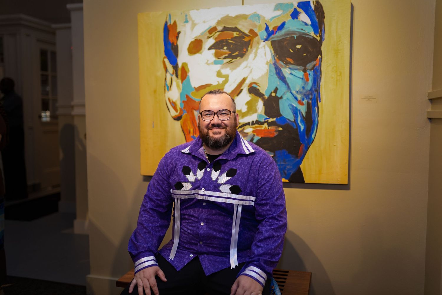 Someone in a purple sweater sits in front of a large, colorful portrait painting with blue and yellow tones in an art gallery.