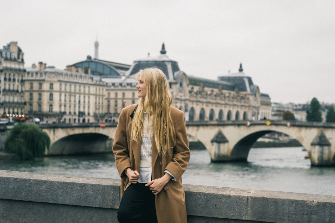 Woman in beige coat standing along Seine River with Musee d'Orsay and historic bridge in Paris background.