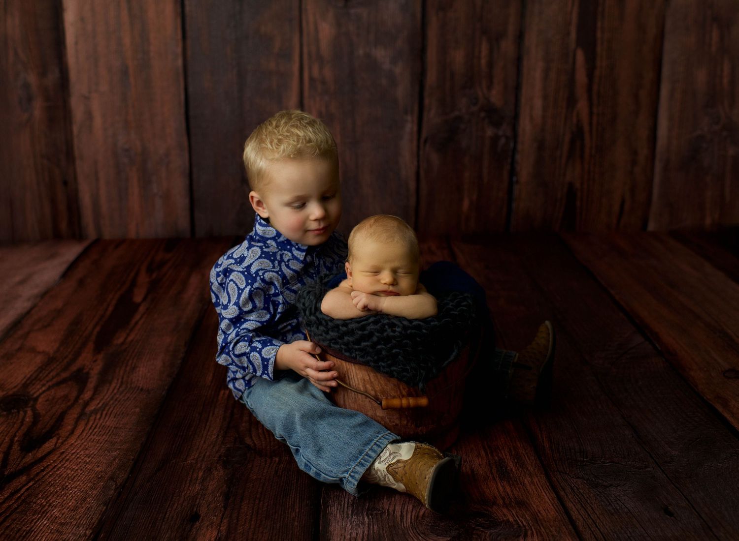 Two young children sit together on a wooden floor against a rustic barn wood wall background.
