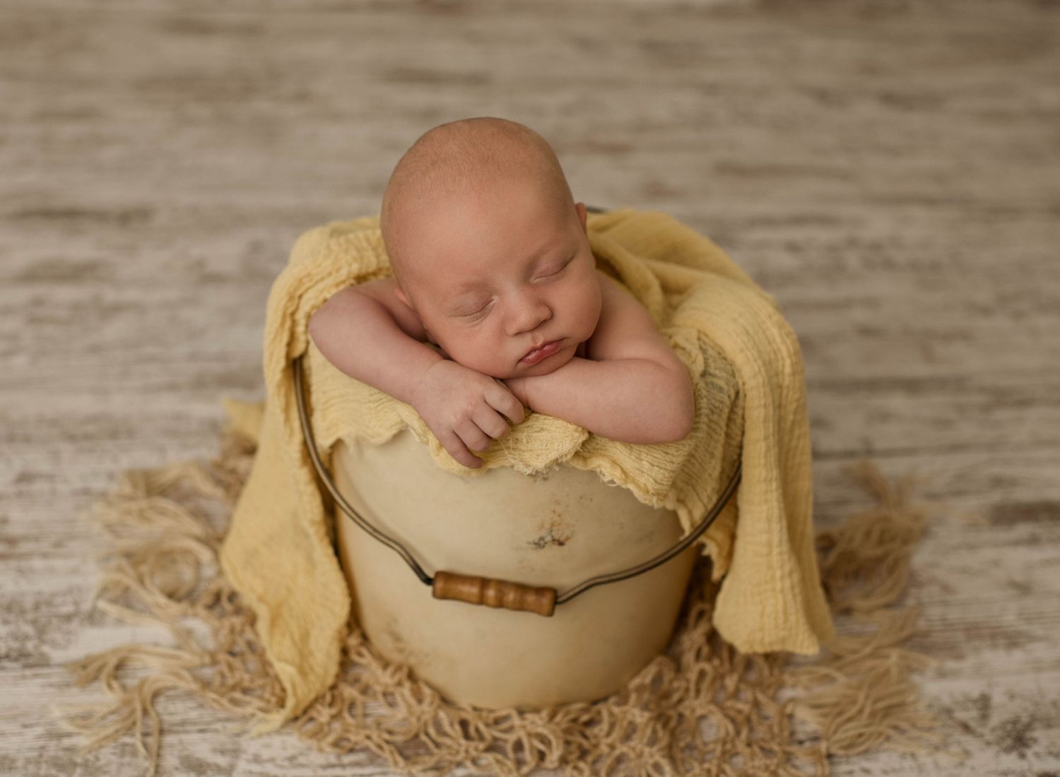 Sleeping newborn wrapped in soft yellow blanket nestles peacefully in rustic wooden bowl on textured floor.