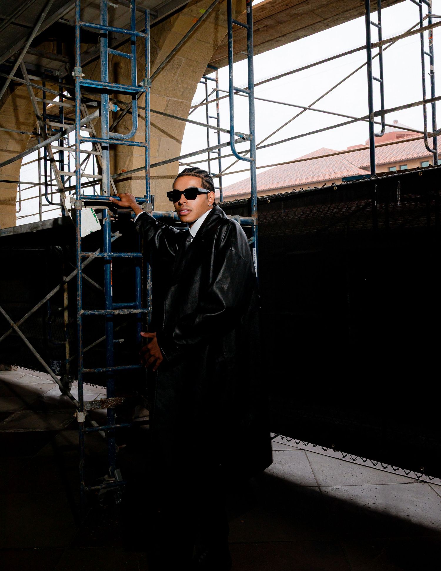 Someone in sunglasses and a black leather jacket poses against scaffolding and industrial architecture in a moody shot.