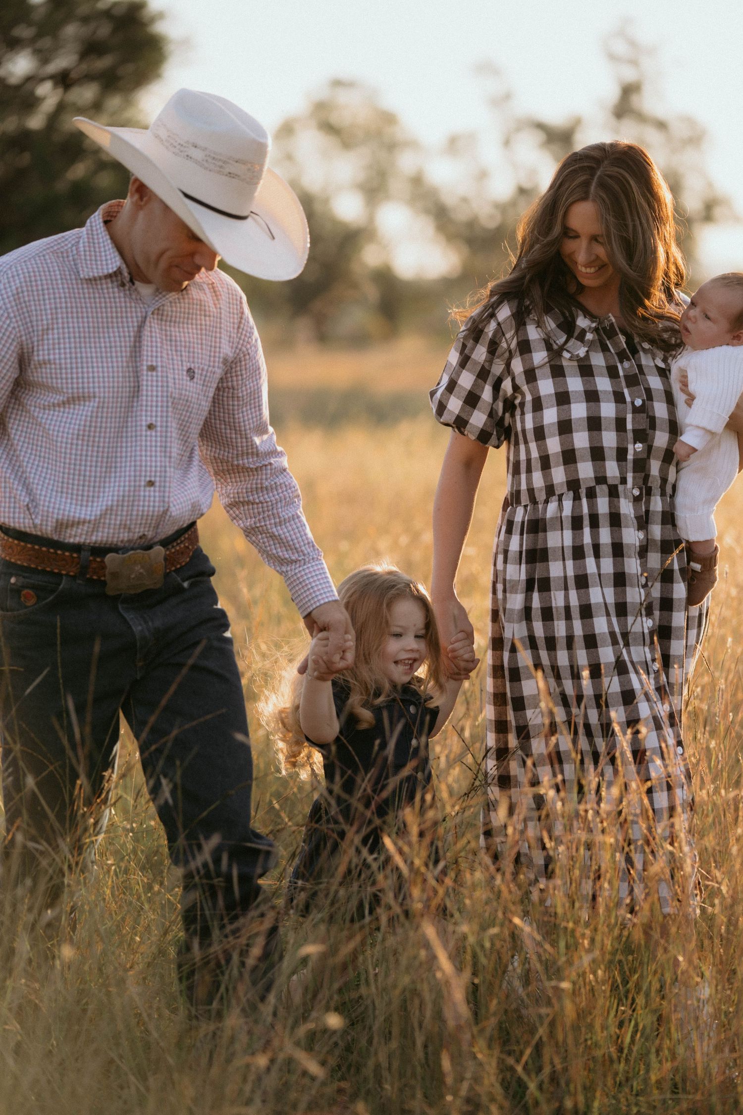 A family in rustic western attire enjoying a tender moment together in a golden sunlit field at sunset.